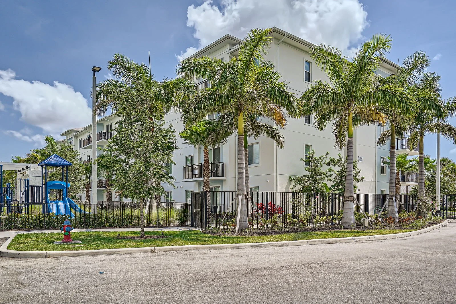 An apartment complex with white walls, surrounded by palm trees, a playground with a blue slide, and a black metal fence. The sky is blue with some clouds.