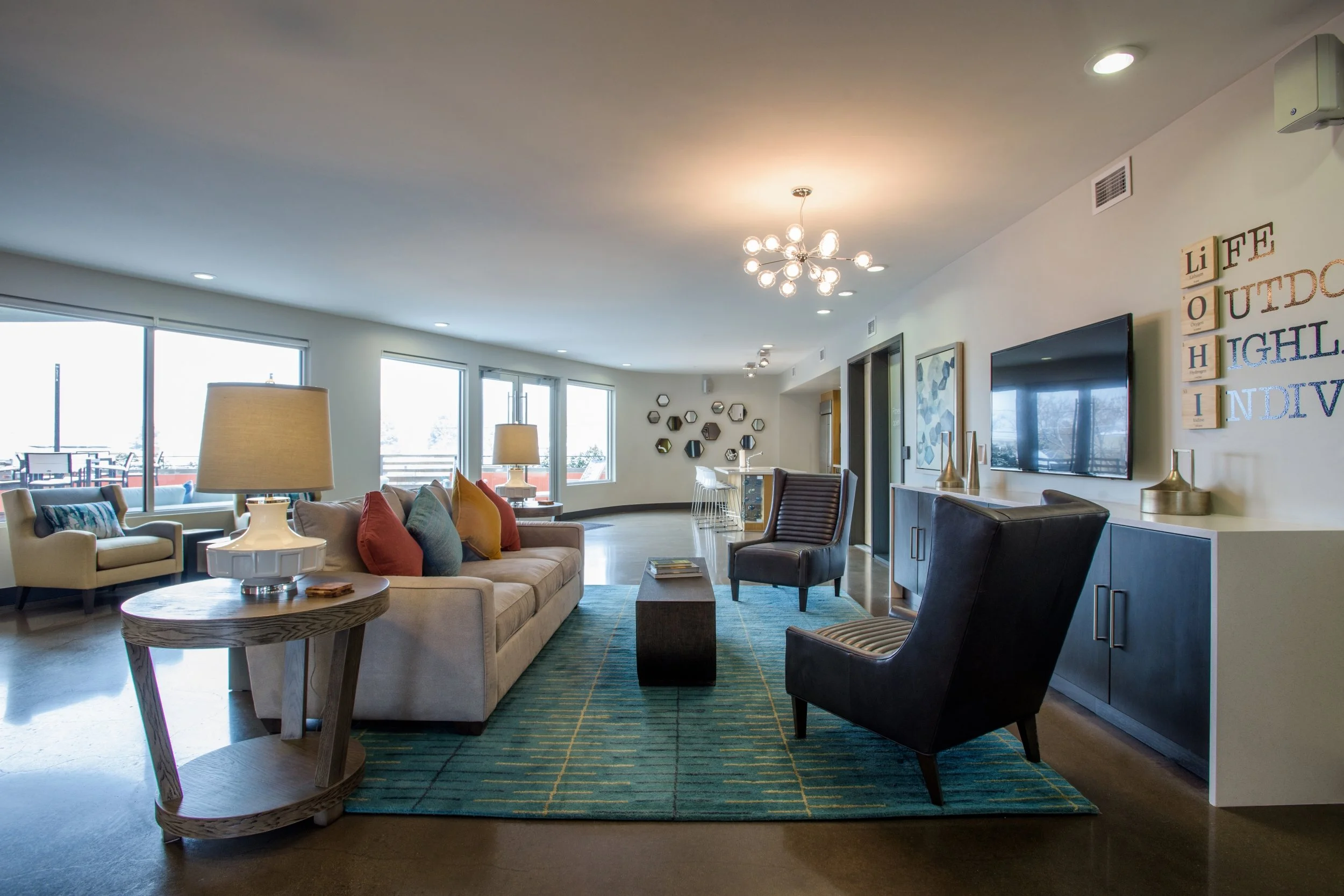 Modern living room with large windows, beige sofa with colorful cushions, two table lamps, black and striped armchairs, a blue and green area rug, wall art, and a flat-screen TV.