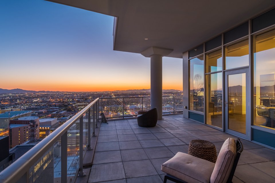 Cityscape view from a high-rise balcony during sunset, with outdoor seating and glass sliding doors.