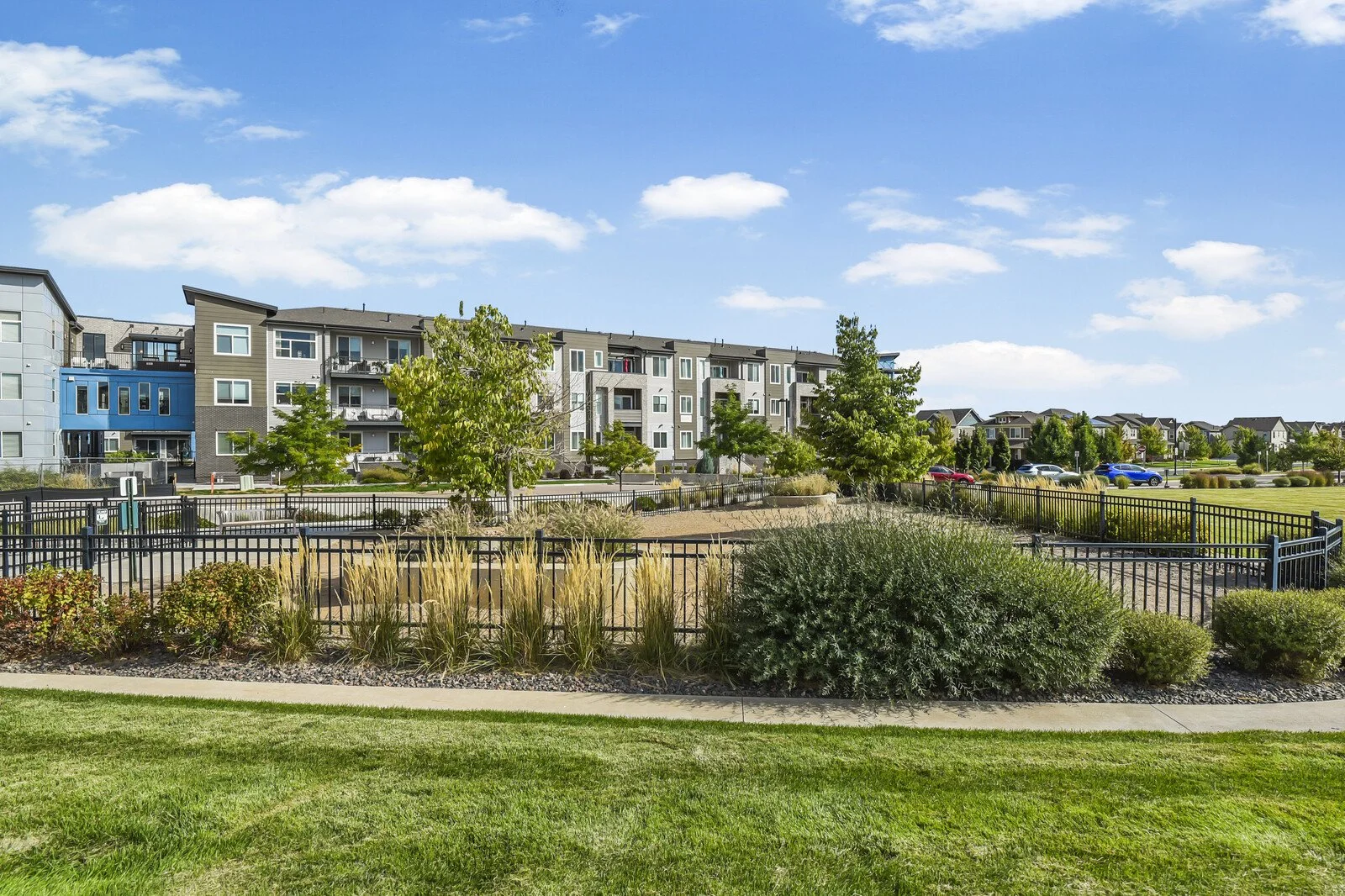 View of a modern apartment complex with landscaped greenery and parking area under a partly cloudy sky.
