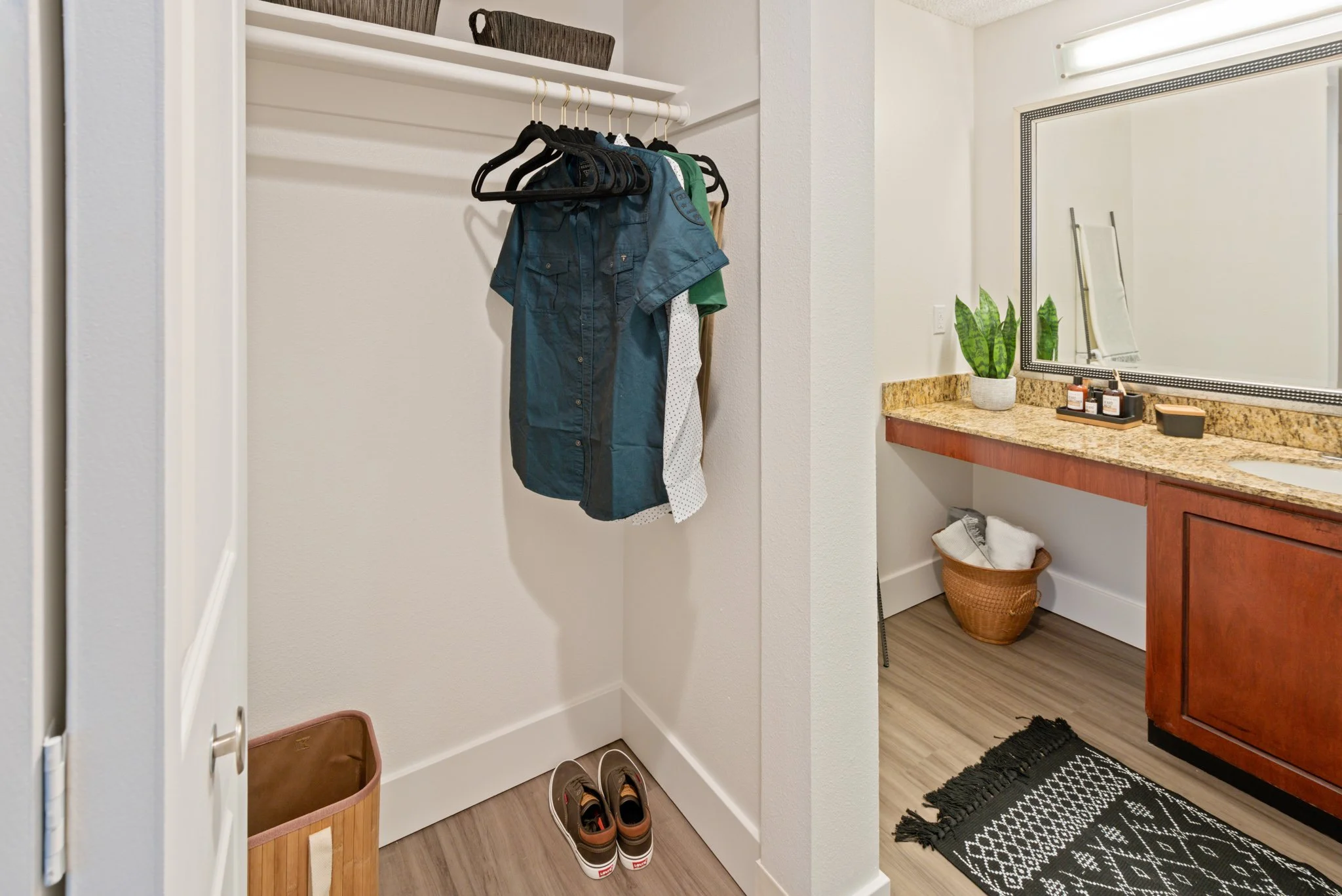 Small closet and bathroom vanity area with a few hanging clothes, shoes on the floor, a basket with clothes, a mirror, a plant, and bathroom essentials on the granite countertop.
