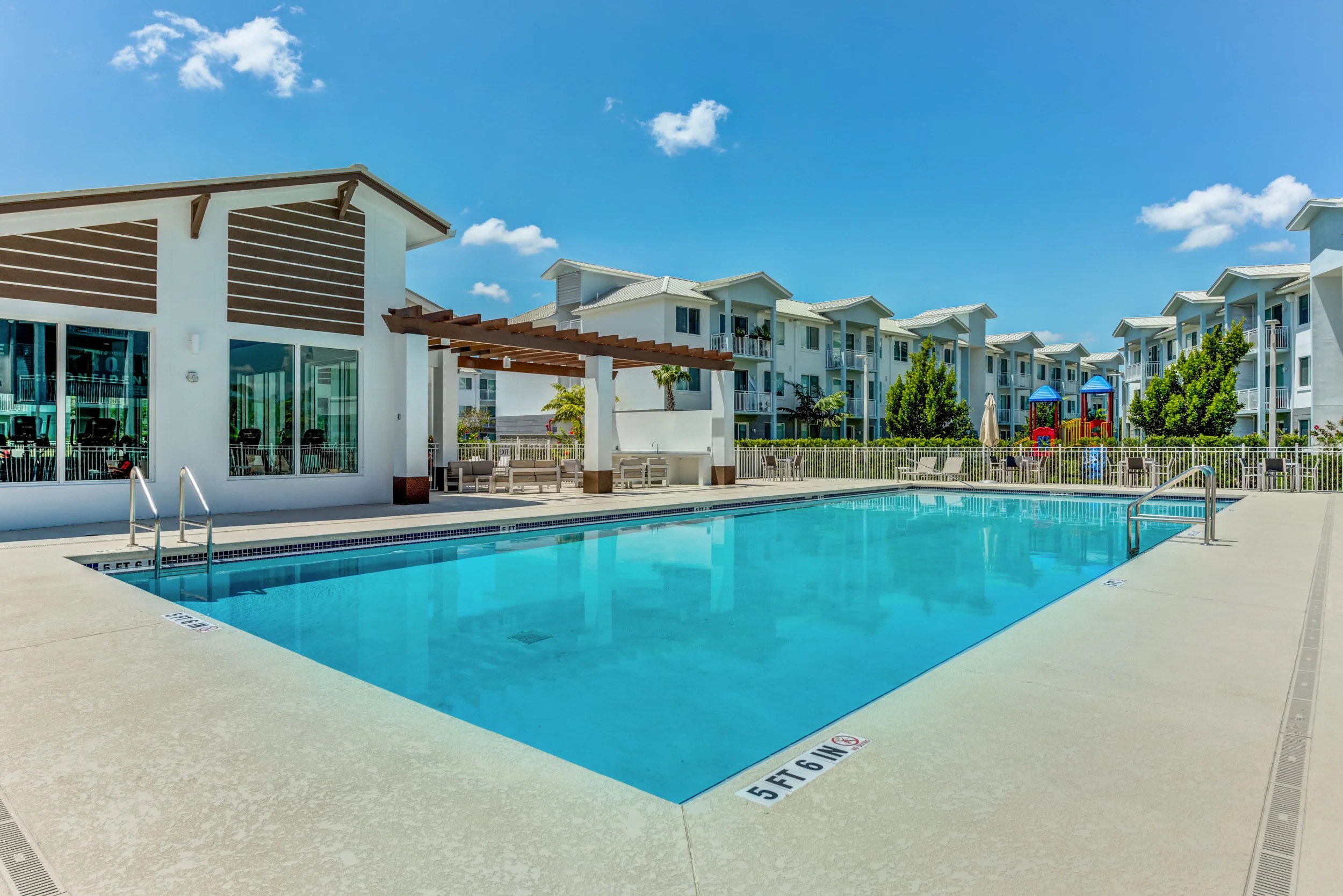 Empty outdoor swimming pool with visible depth markers, surrounded by lounge chairs and colorful playground equipment, residential buildings in the background under a partly cloudy sky.