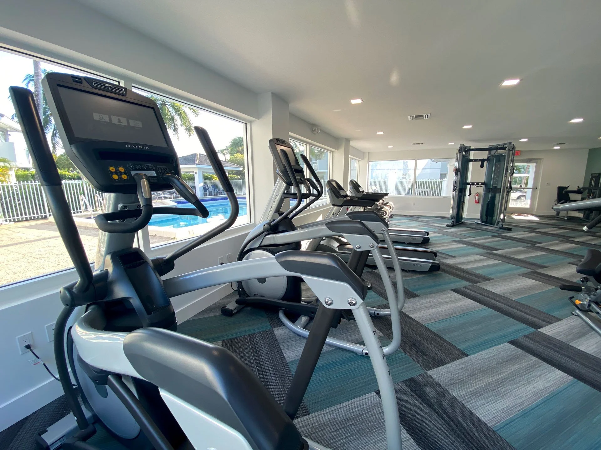 Gym room with exercise equipment including elliptical machines, treadmills, and weights, with large windows showing outdoor pool area.
