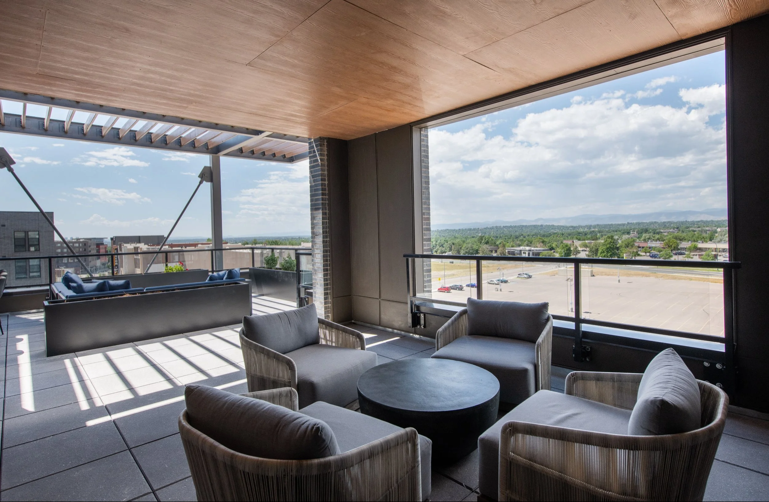 Indoor lounge area with armchairs and a round coffee table, large windows offering a city and landscape view, some patio furniture, blue sky with clouds.