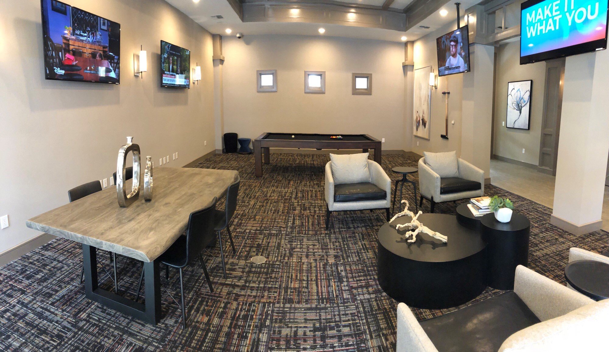A modern lounge area with multiple wall-mounted television screens, a pool table in the center, comfortable armchairs, side tables, decorative sculptures, and artwork on the walls. There are small windows and bright lighting.