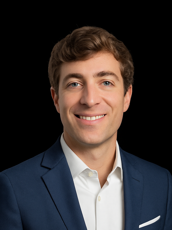 A young man with brown hair and blue eyes, wearing a dark blazer over a collared shirt, smiling against a dark blurred background.