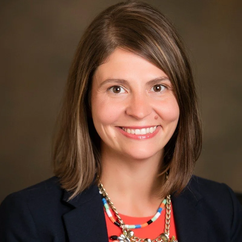 Headshot of a woman with shoulder-length brown hair, smiling, wearing a black blazer, colorful necklace, and a red top.