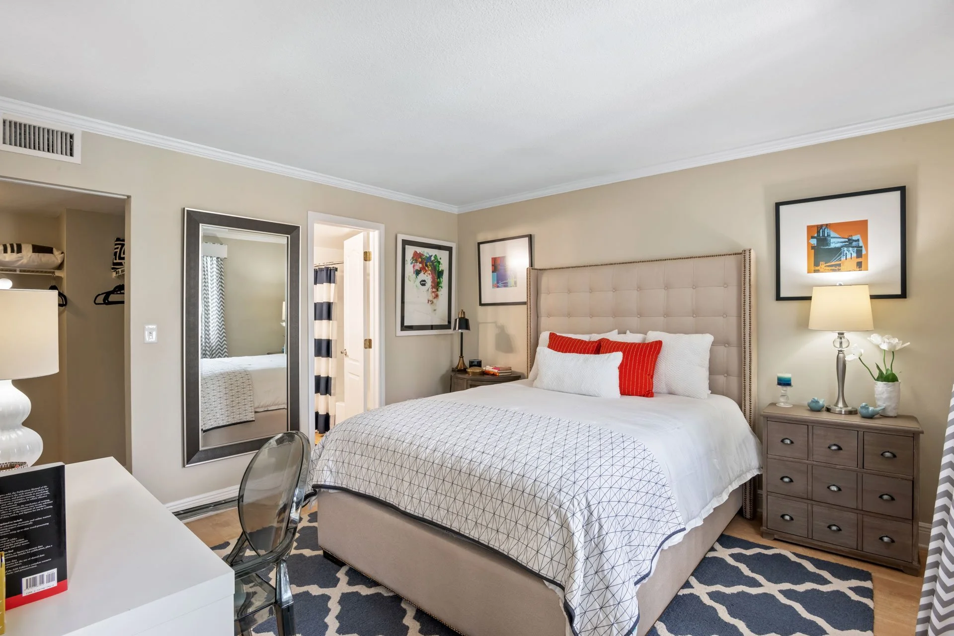 A bedroom with a beige upholstered bed with white and red pillows, light-colored dresser, framed artwork on beige walls, a mirror, and a patterned rug.