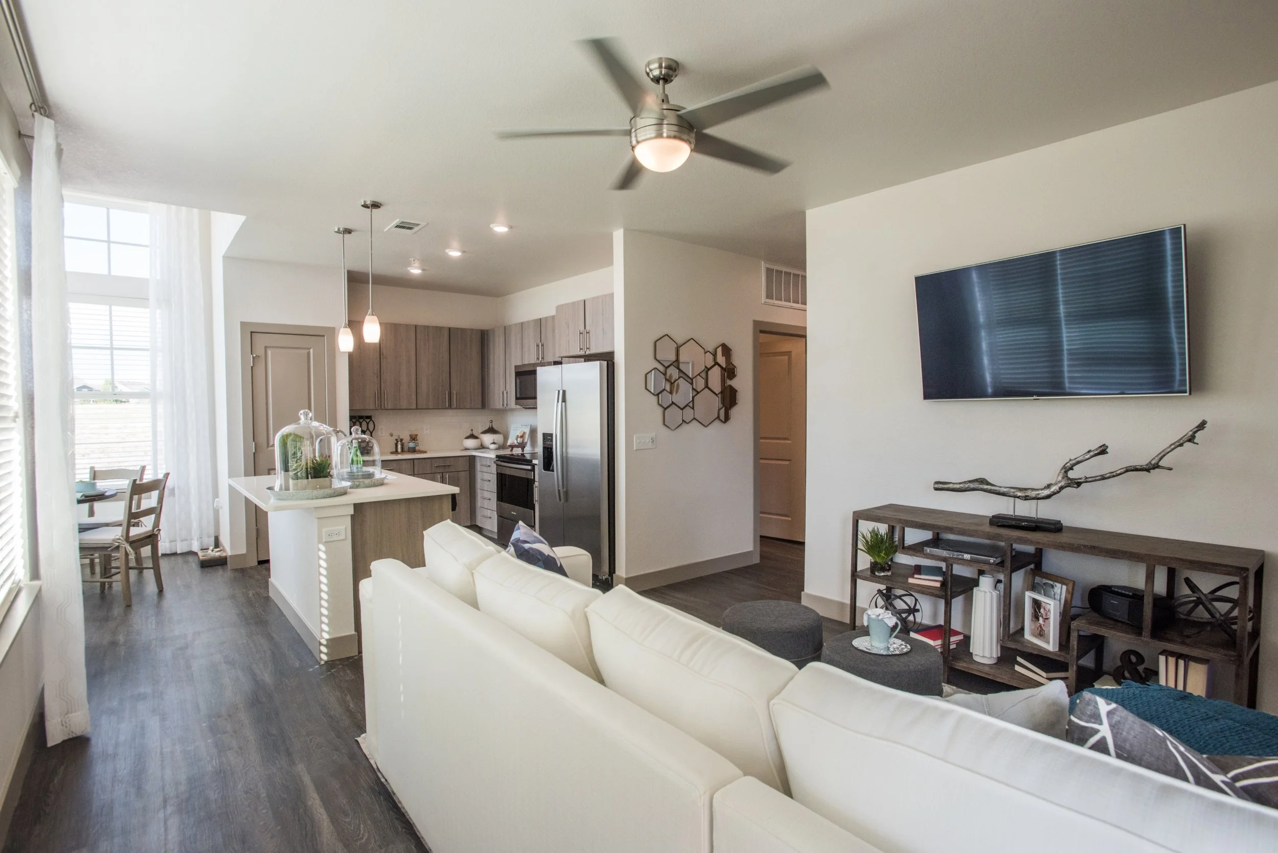 Open-concept living room and kitchen with white sofa, wall-mounted TV, and modern decor in a bright space.