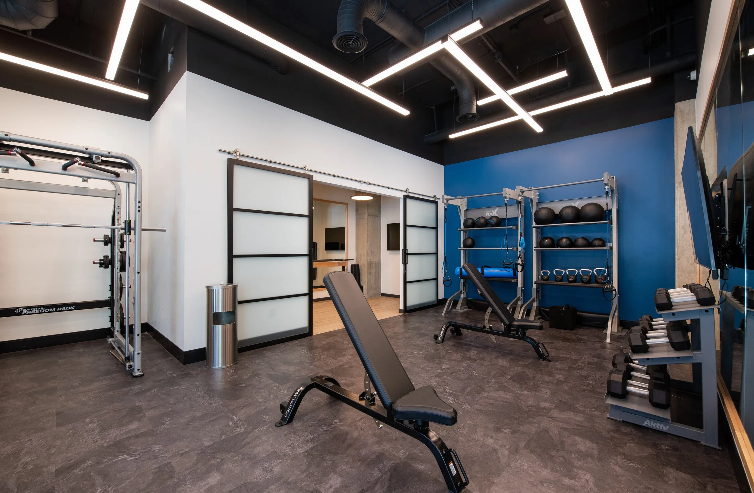Modern gym with workout equipment including a weight rack, adjustable benches, medicine balls, kettlebells, and dumbbells, with a black and white color scheme and bright overhead lighting.