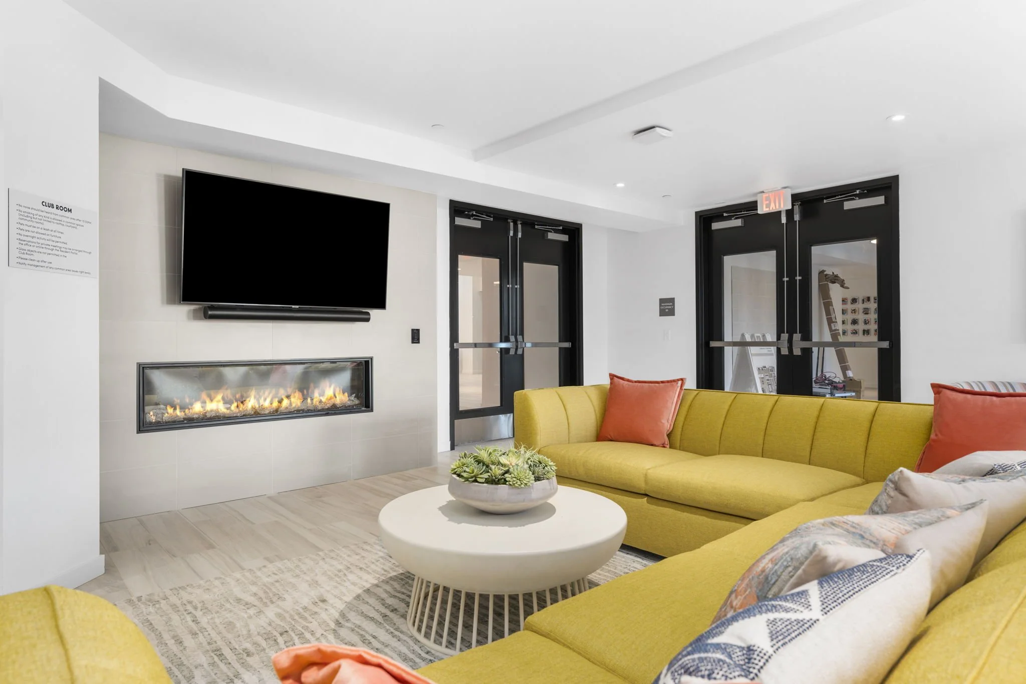 Modern lounge with yellow sectional sofa, orange and patterned throw pillows, white round coffee table with succulent plant, wall-mounted flat-screen TV, and electric fireplace, black-framed glass doors leading outside, and white walls and ceiling.