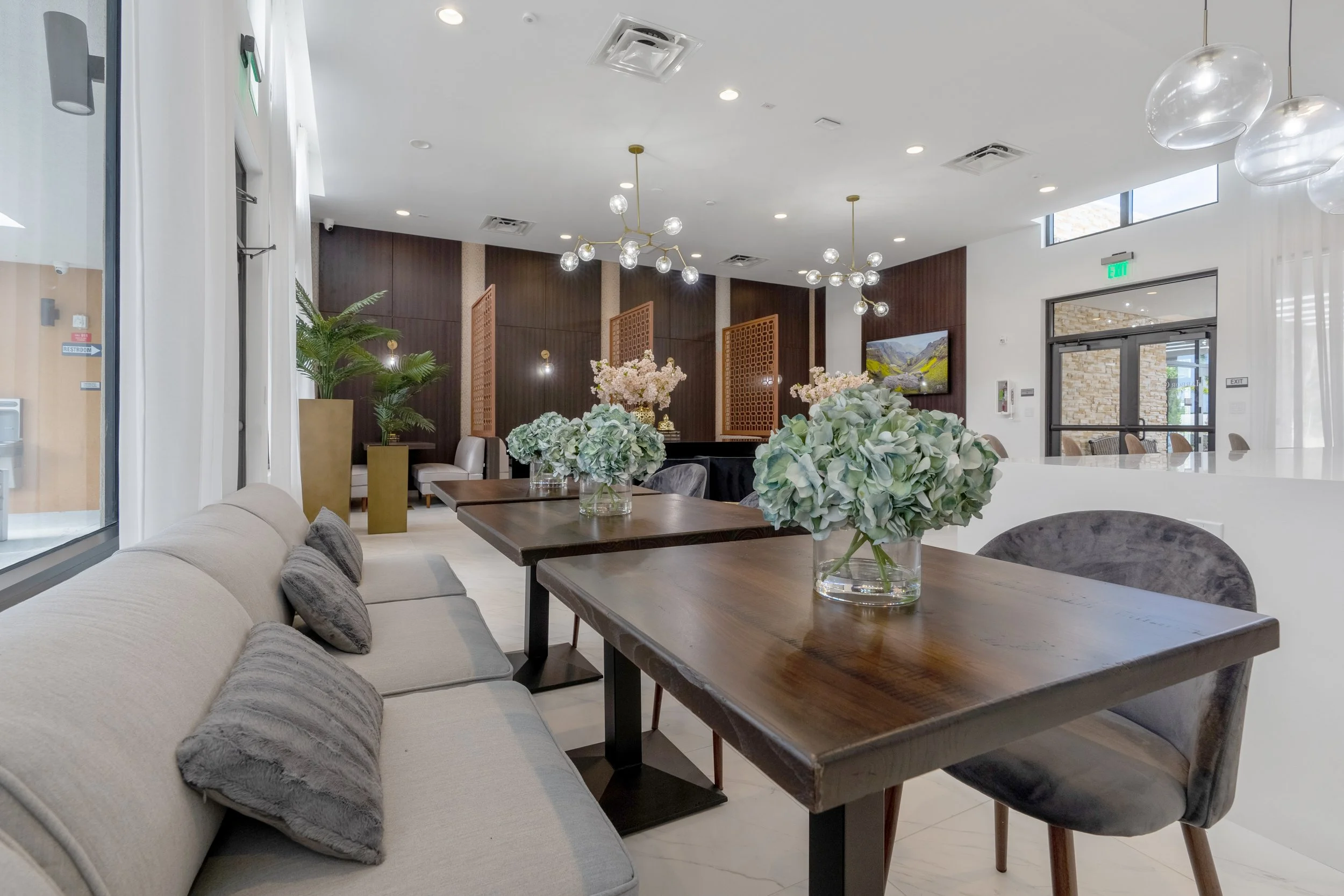 Elegant restaurant interior with beige sofa and dark wooden table adorned with glass vases of white and green hydrangeas, modern hanging pendant lights, a dark wood accent wall, and large windows letting in natural light.