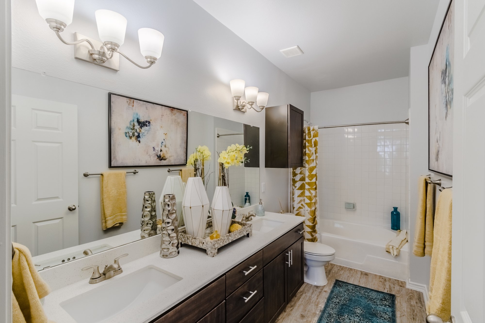 Bathroom with double sink vanity, decorative vases, a large mirror, artwork, yellow towels, a bathtub with a yellow and white curtain, and a blue rug.