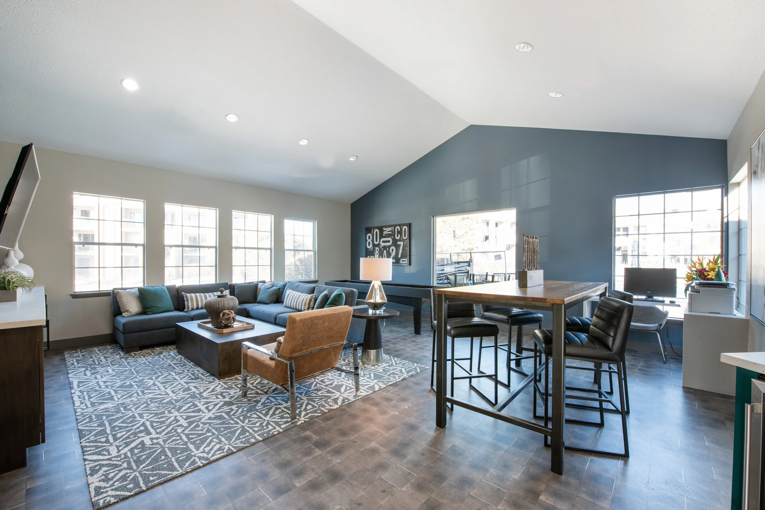 Open-concept living room and dining area in modern apartment with large windows, gray wall, sectional sofa, coffee table, armchair, bar-height table with stools, and workspace with computer.