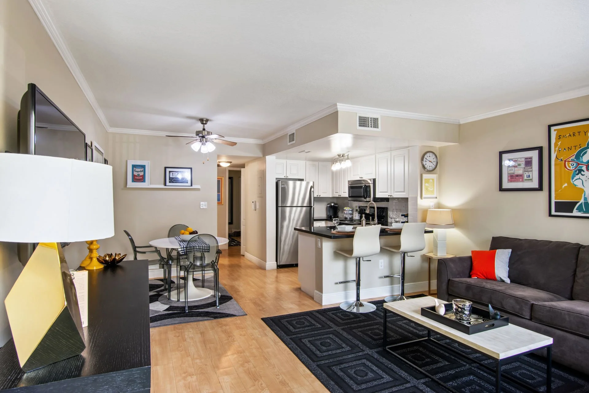 A living room and kitchen with hardwood floors, a gray couch with colorful pillows, a modern coffee table, a dining table with chairs, a kitchen breakfast bar with stools, stainless steel appliances, and wall art.