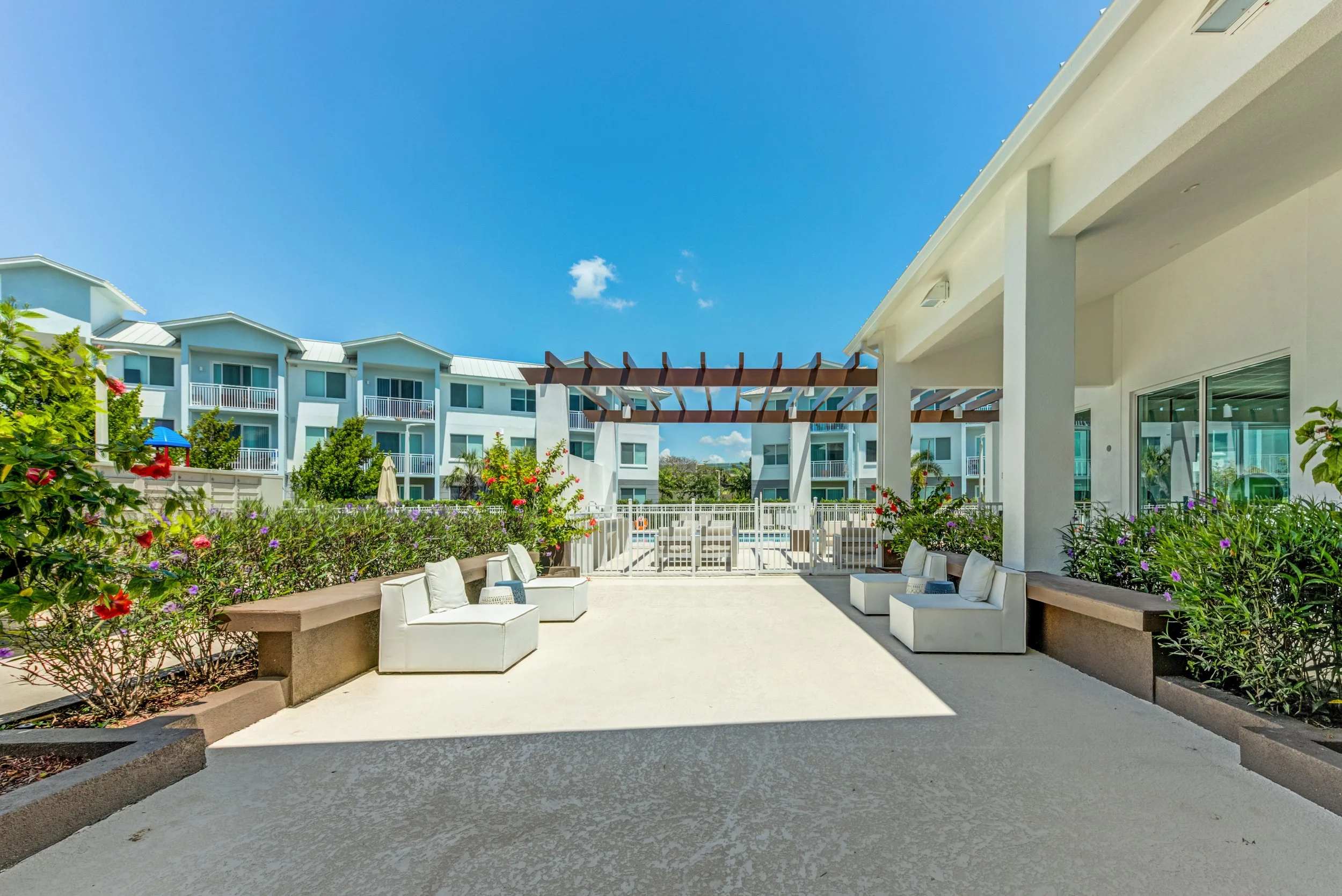 Outdoor patio area with white lounge chairs, benches, and lush flowering bushes under a bright blue sky, part of a modern residential complex.