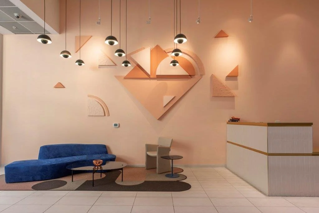 Modern hotel lobby with abstract geometric wall art, blue sofa, beige chairs, two tables, and a white reception desk.