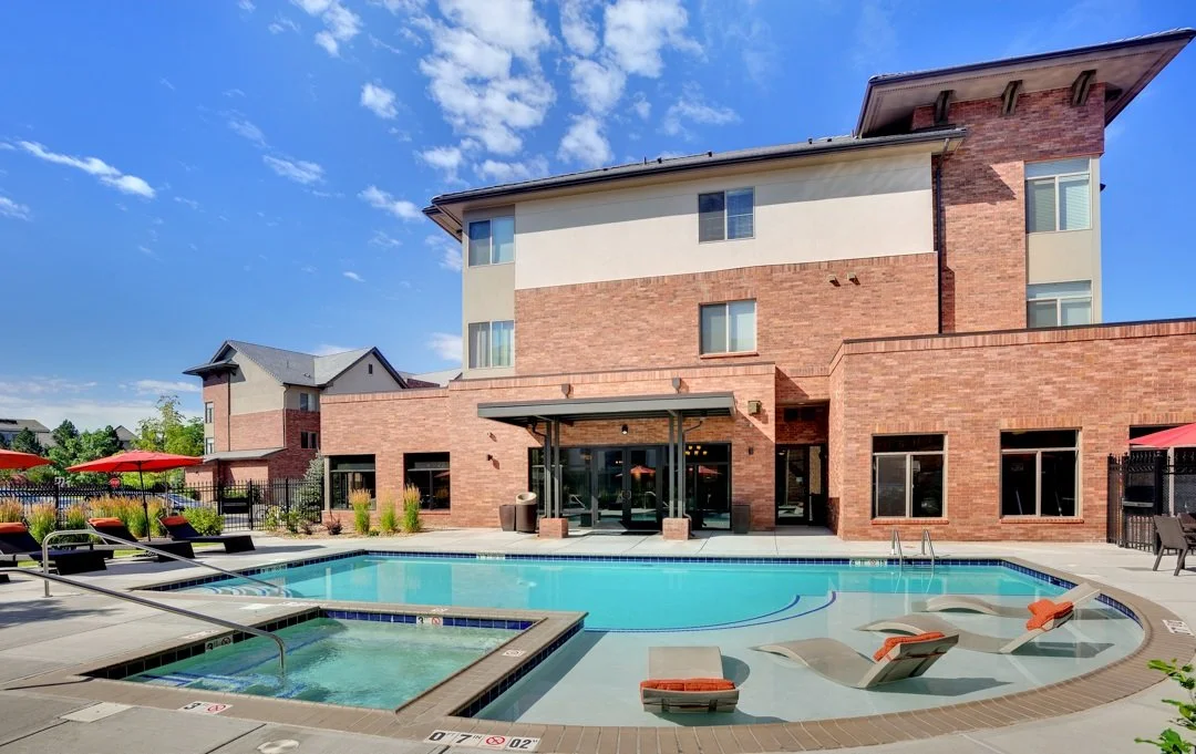 An outdoor swimming pool area with lounge chairs, umbrellas, and a hot tub, in front of a multi-story brick and stucco residential building on a sunny day.