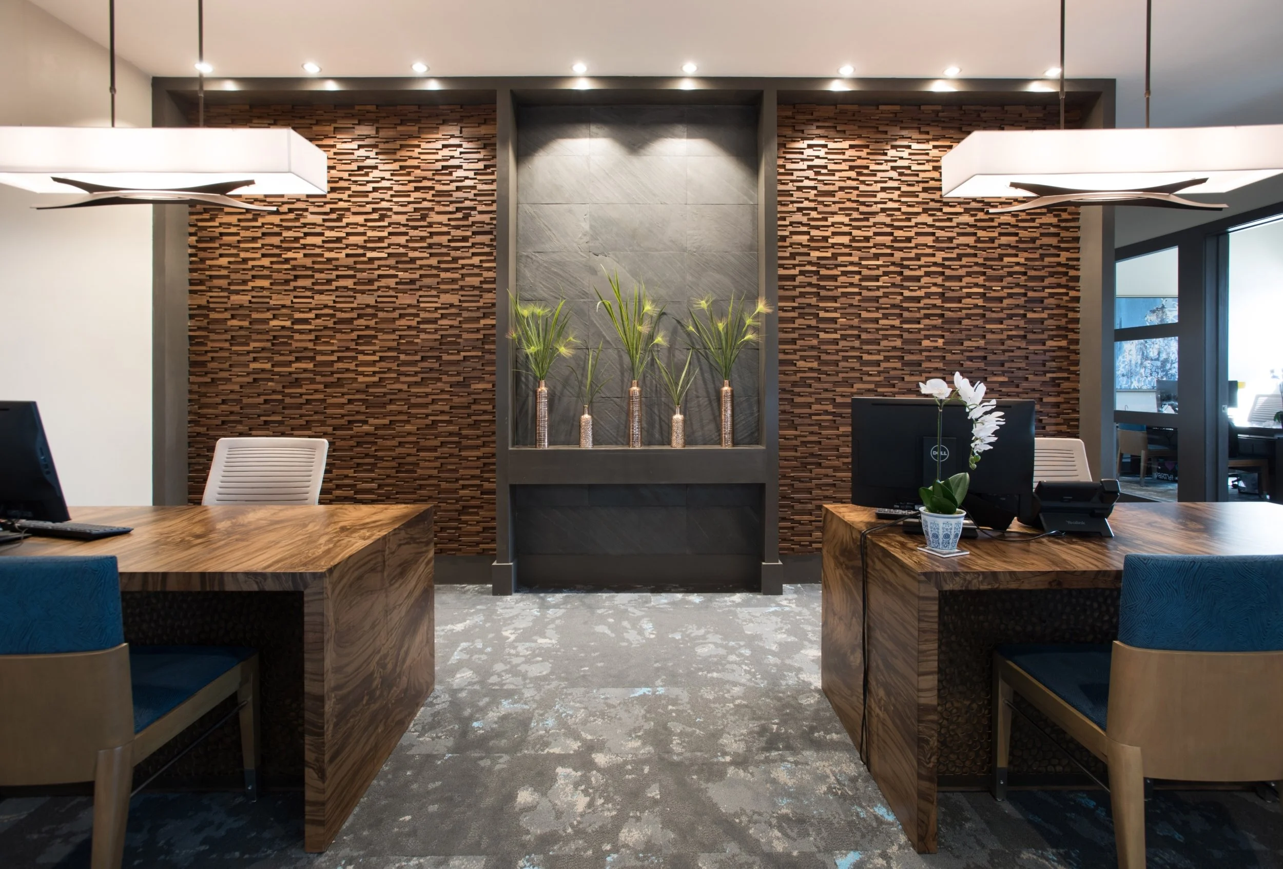 Modern office reception area with two wooden desks, a computer and a phone on one, a desk with a potted orchid, a white chair, and a wall with decorative stone and wood design, along with multiple potted plants and eclectic lighting fixtures.