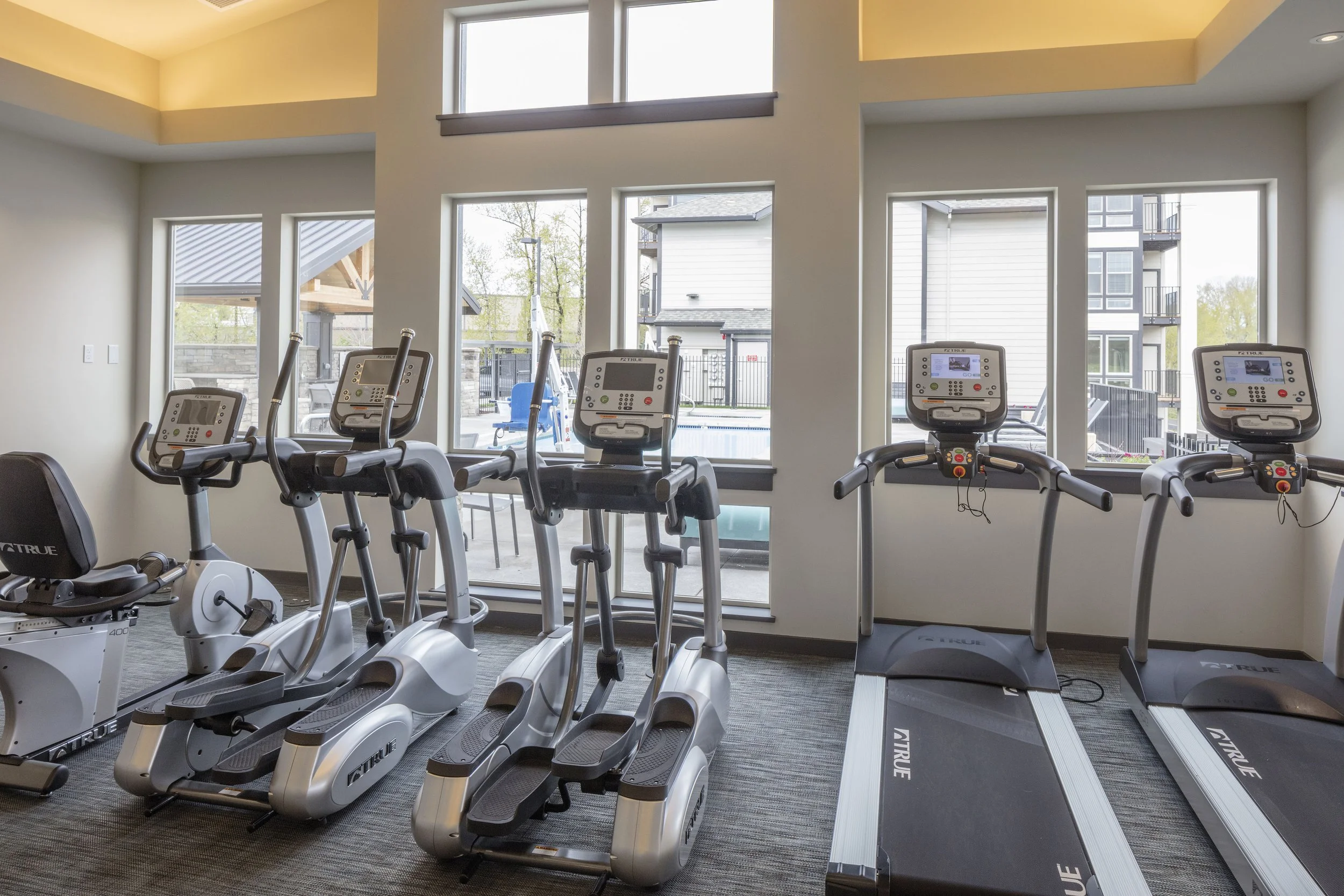 Indoor gym with exercise equipment including elliptical machines and treadmills facing large windows with outdoor view.