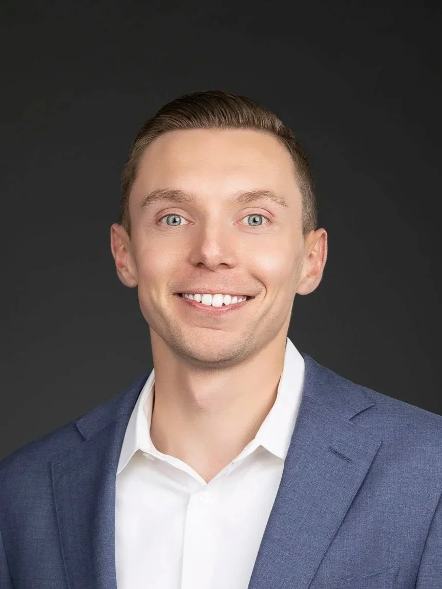 A young man with light skin, short light brown hair, blue eyes, wearing a blue suit jacket and white shirt, smiling in front of a dark gray background.