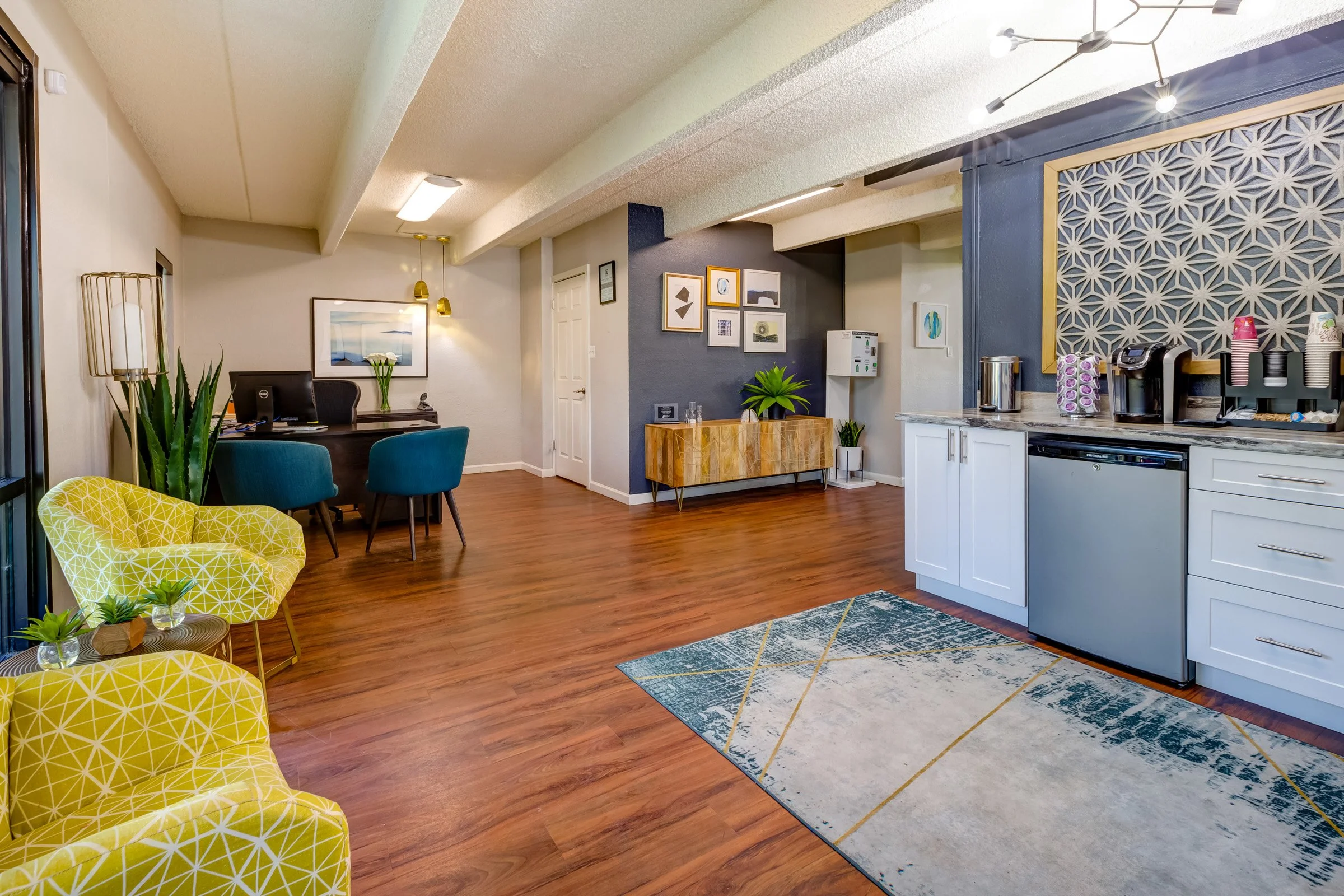Interior of a modern apartment with hardwood floors, a kitchenette with white cabinets and a countertop, a small rug, a yellow patterned armchair and matching side tables, a desk with a computer and chairs, a decorative wall with framed artwork, and 