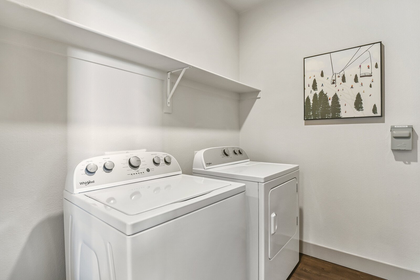A laundry room with a white Whirlpool washing machine and a matching white dryer, a white shelf above them, a framed picture on the wall, and a wall-mounted utility box.