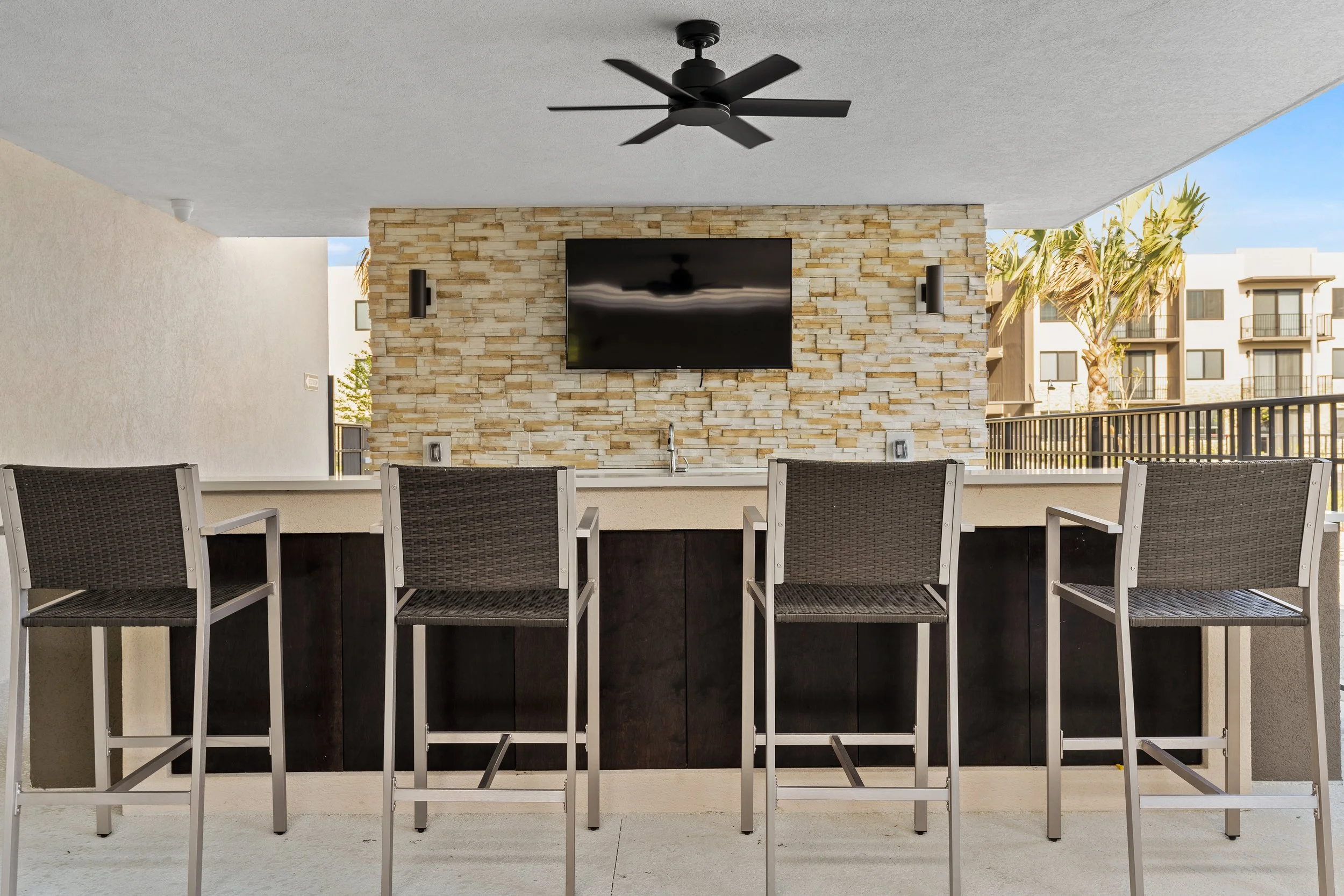 Outdoor balcony with a bar area, high chairs, a stone wall with a mounted flat-screen TV, ceiling fan, and windows showing a sunny day with palm trees.
