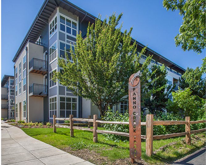 Multi-story apartment building with large windows, surrounded by trees, a wooden fence, and a sign for Fanno Creek Trail.