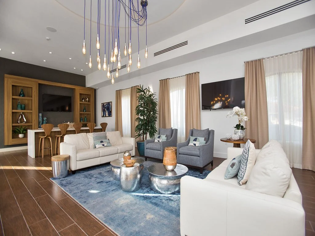 Contemporary living room with white and grey sofas, glass and metallic accent tables, a blue area rug, and a large wooden entertainment center with built-in shelves. Features a flat-screen TV, tall curtains, potted plant, and modern chandelier.