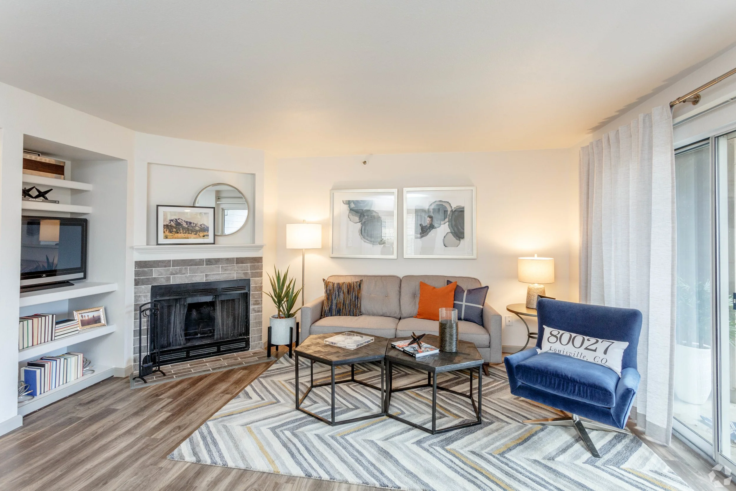 Living room with a beige sofa, blue armchair, coffee tables, fireplace, wall art, and a glass sliding door with curtains.