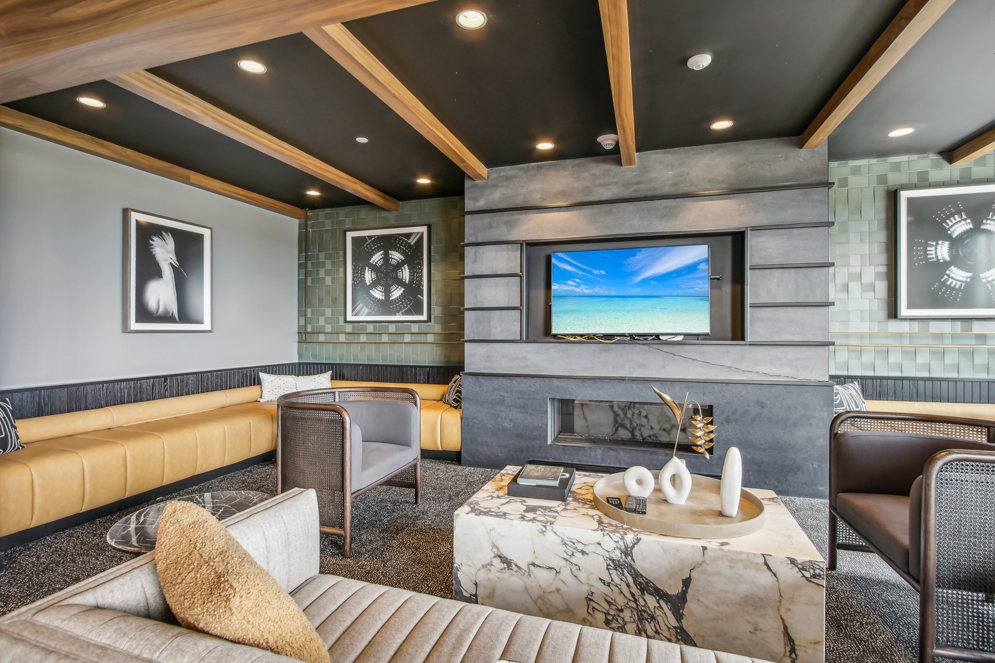 Modern hotel lobby with a stone fireplace, TV displaying a beach scene, and seating areas including a beige sofa, gray armchair, and brown and yellow benches. Artworks on the wall and decorative vases on a marble coffee table.