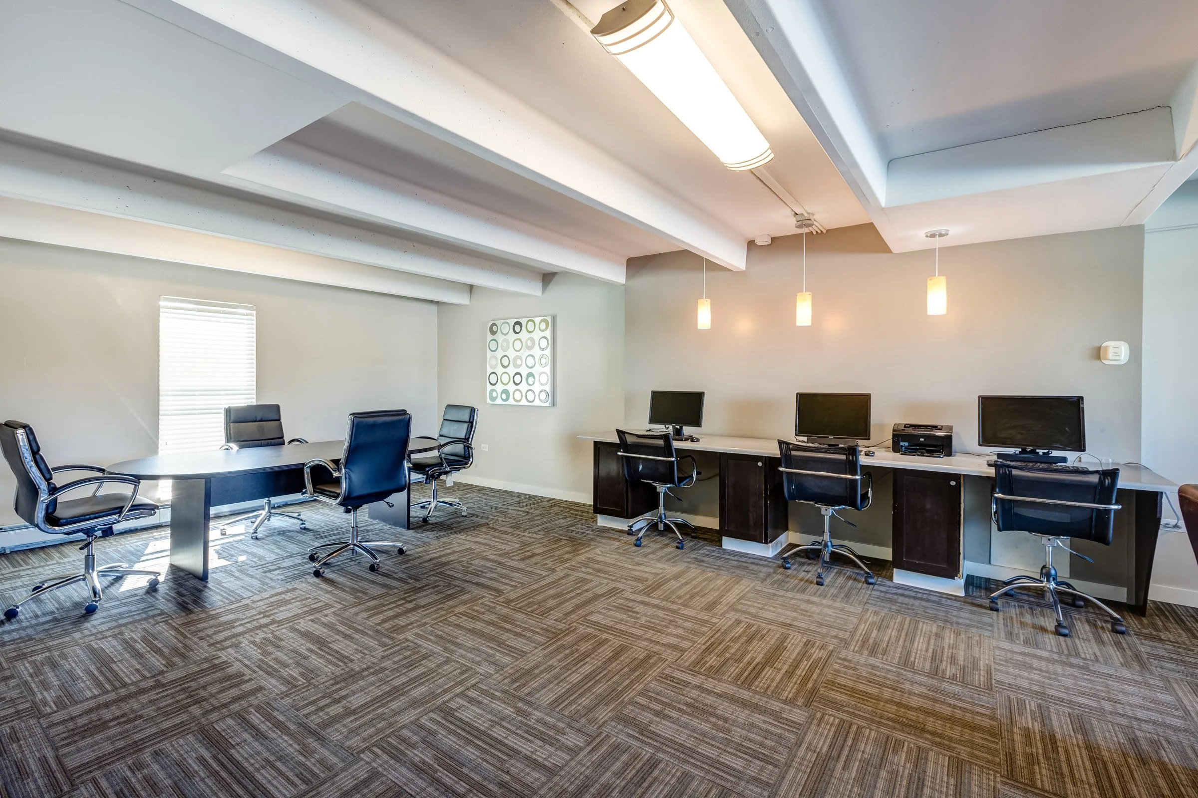 An empty modern office conference room with a large oval table, six black office chairs, and a row of three computer workstations along the wall. The room has beige walls, a patterned carpet, and ceiling lighting.