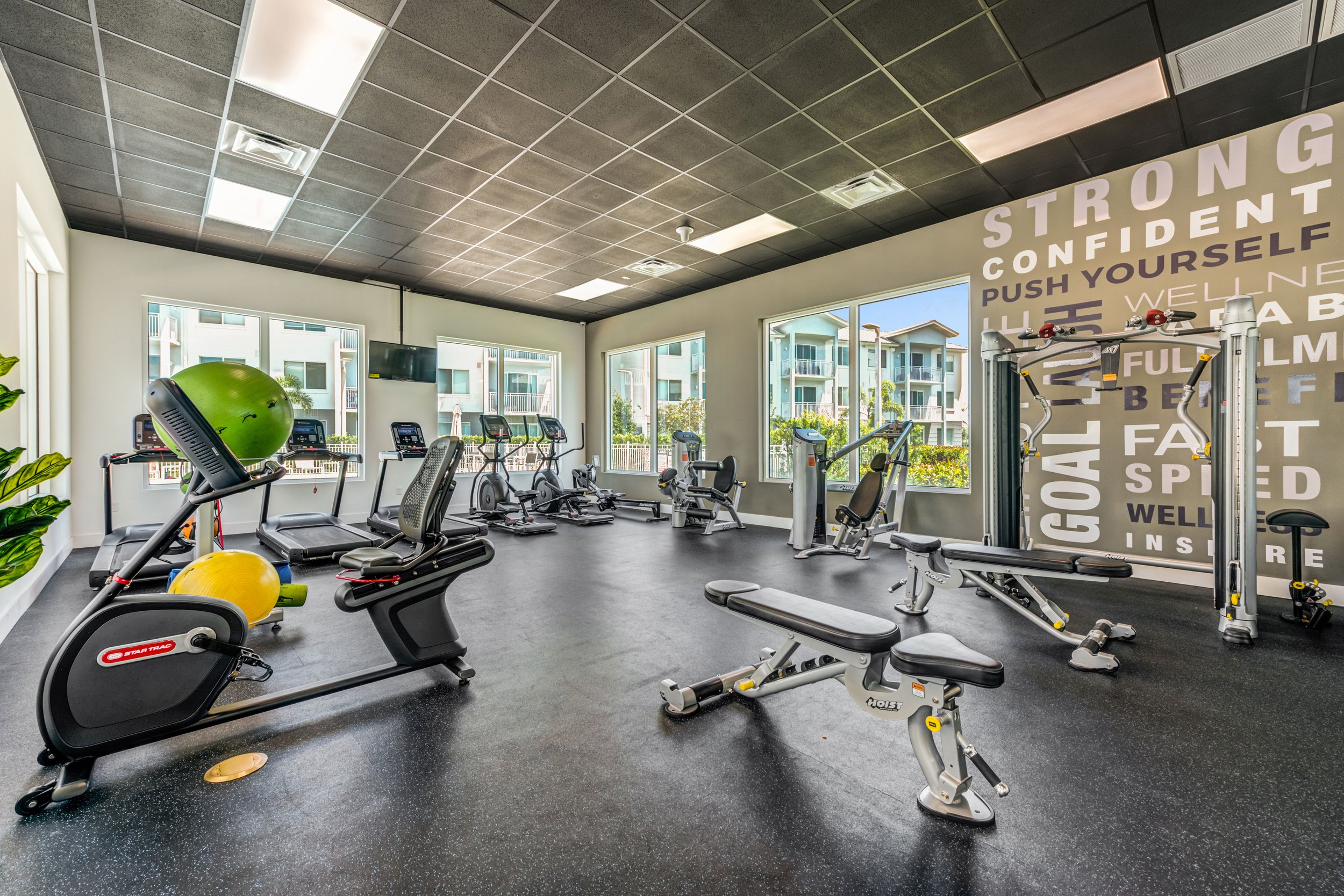 A well-lit gym with various exercise equipment, large windows, and motivational wall art.