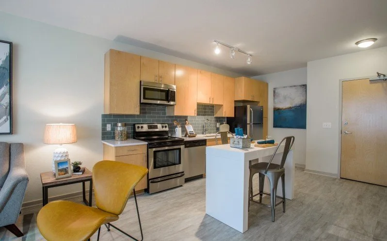 Modern kitchen with wooden cabinets, stainless steel appliances, gray tile backsplash, small island, and a seating area with a yellow chair.