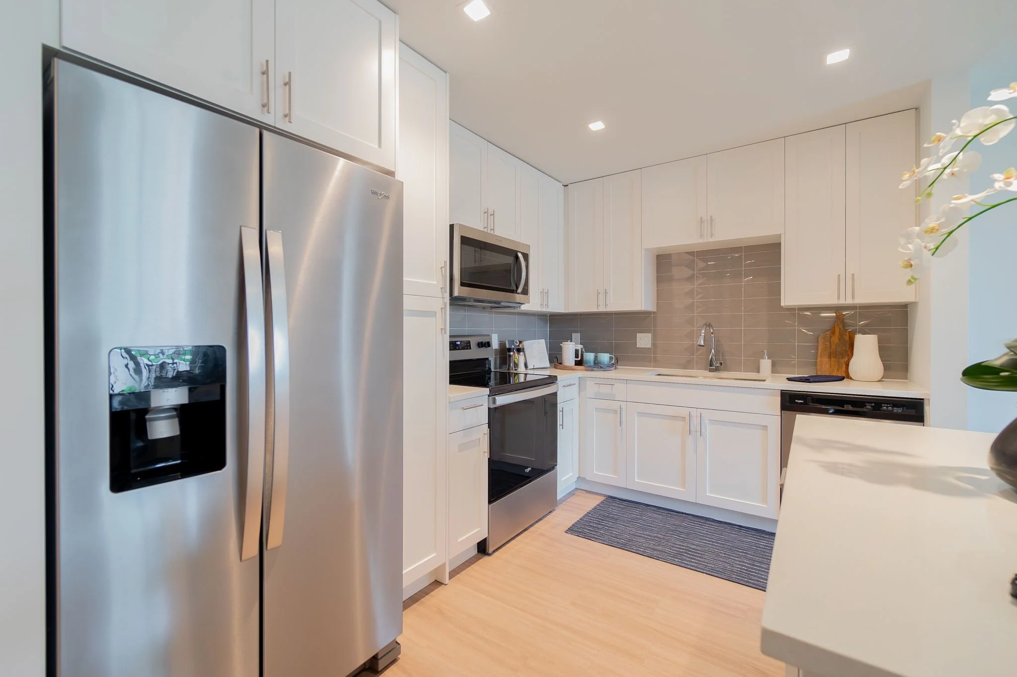 Modern kitchen with white cabinets, stainless steel refrigerator, microwave, oven, and dishwasher, beige tile backsplash, and light wood flooring.