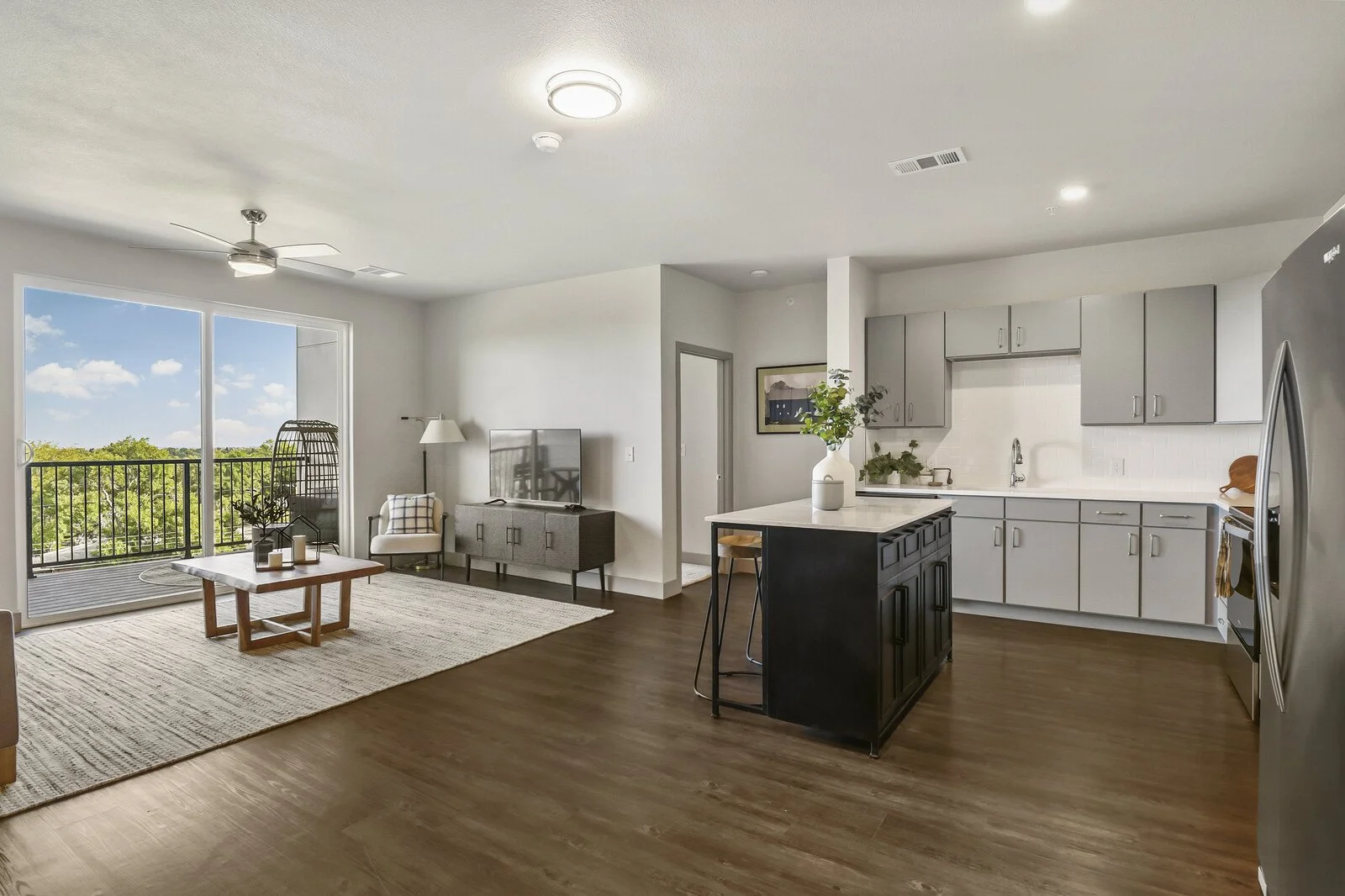Living room and kitchen area in an apartment with wooden floor, sliding glass door leading to balcony, modern furniture, and a kitchen island with plants.