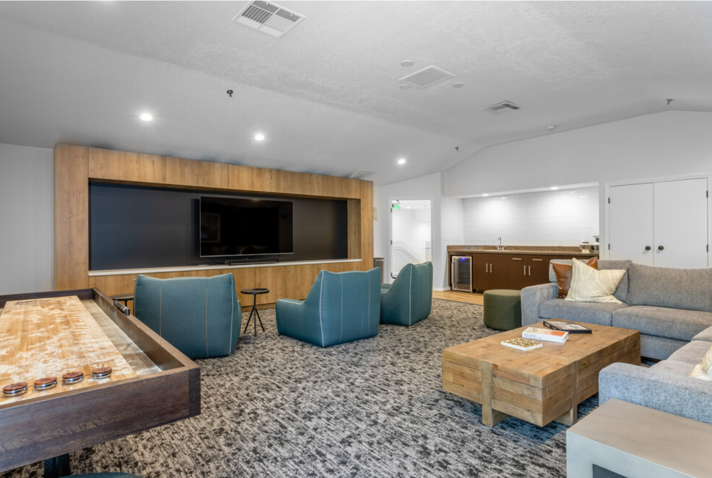 Modern lounge area with a large flat-screen TV on a wooden accent wall, teal armchairs, gray sectional sofa with pillows, wooden coffee table, game table, and a small kitchenette in the background.