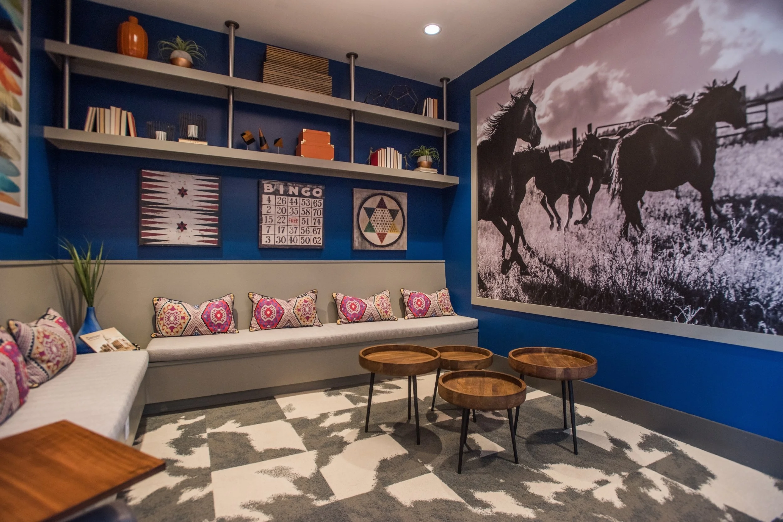 A cozy living room with a blue accent wall, a large black-and-white mural of running horses, a long bench with colorful patterned pillows, three wooden coffee tables, a patterned gray and white rug, and decorative shelves with books, plants, and art 