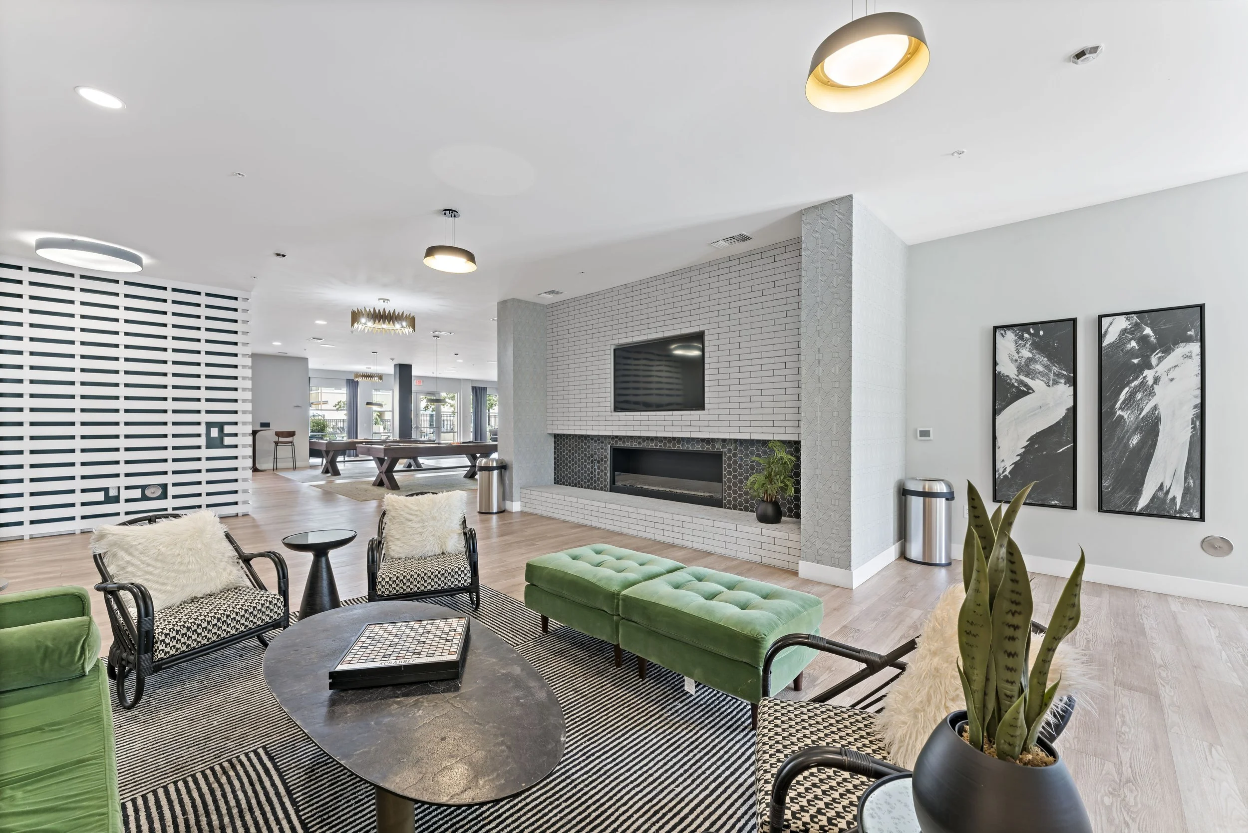Modern living room with white brick fireplace, wall-mounted TV, black and white abstract art, green velvet bench, patterned chairs with fluffy cushions, black oval coffee table, potted snake plant, light wood flooring, and ceiling lights.