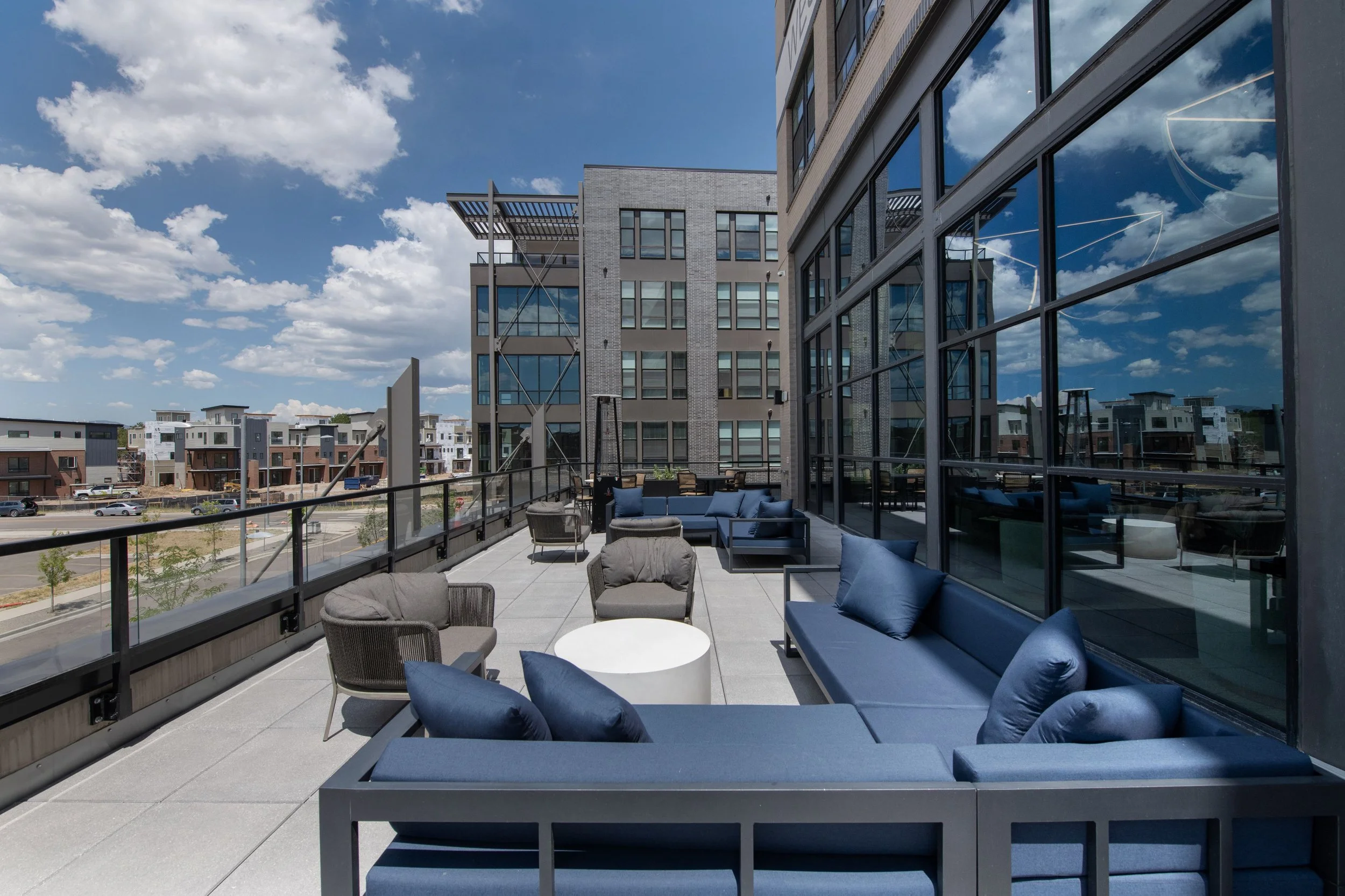 Outdoor rooftop balcony with modern blue and gray seating, cushions, and tables, overlooking an urban area with apartment buildings under a partly cloudy sky.