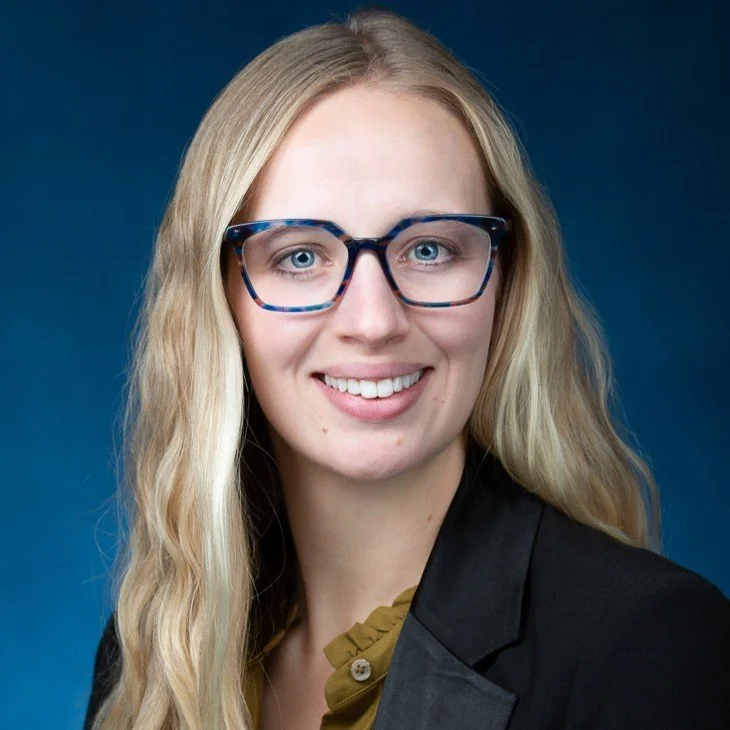 A woman with long blonde hair, wearing blue glasses and a black blazer, smiling against a blue background.
