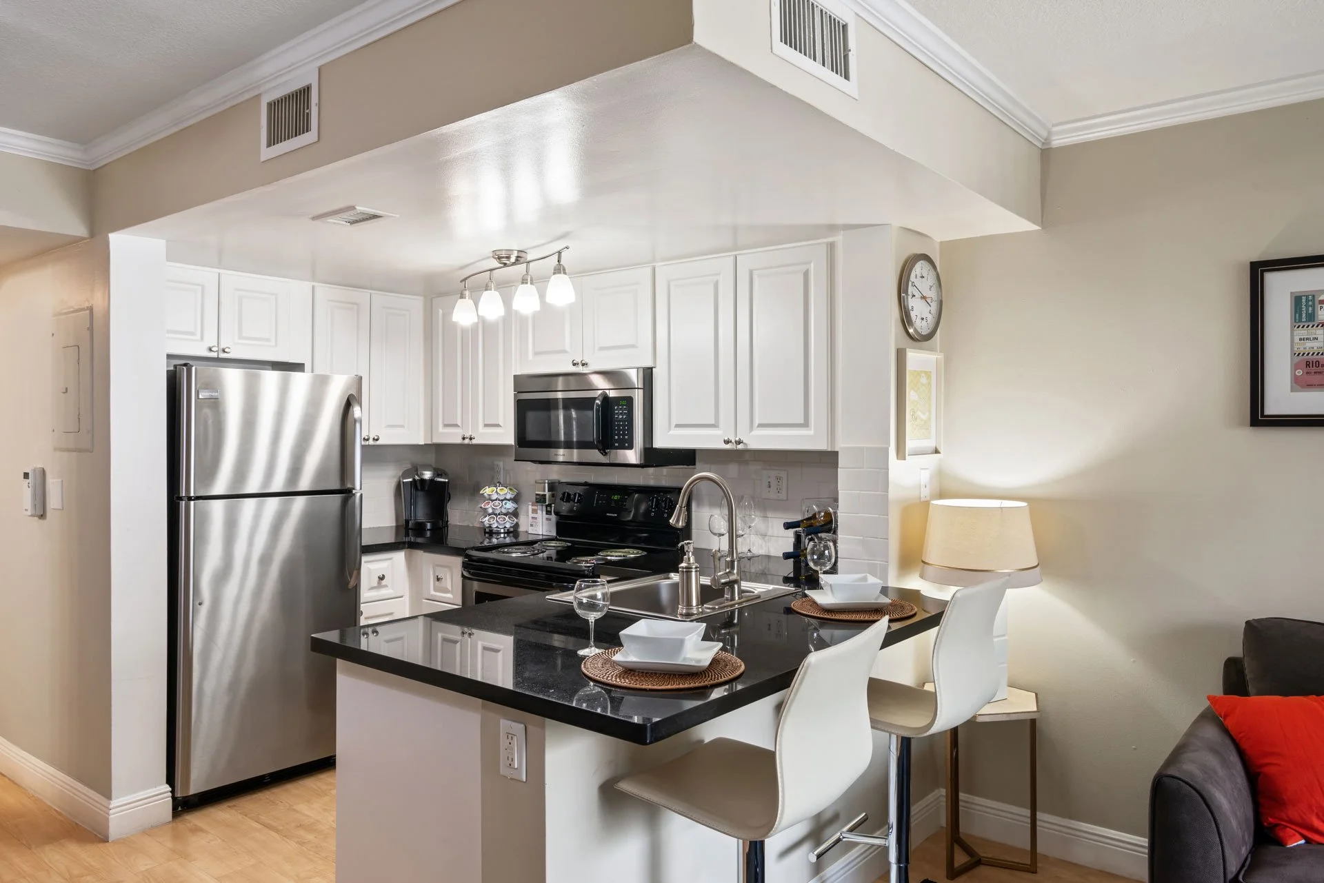 Modern kitchen with white cabinets, stainless steel refrigerator, microwave, and oven, black countertop island with place settings, two white bar stools, a small table with a lamp, and wall art.