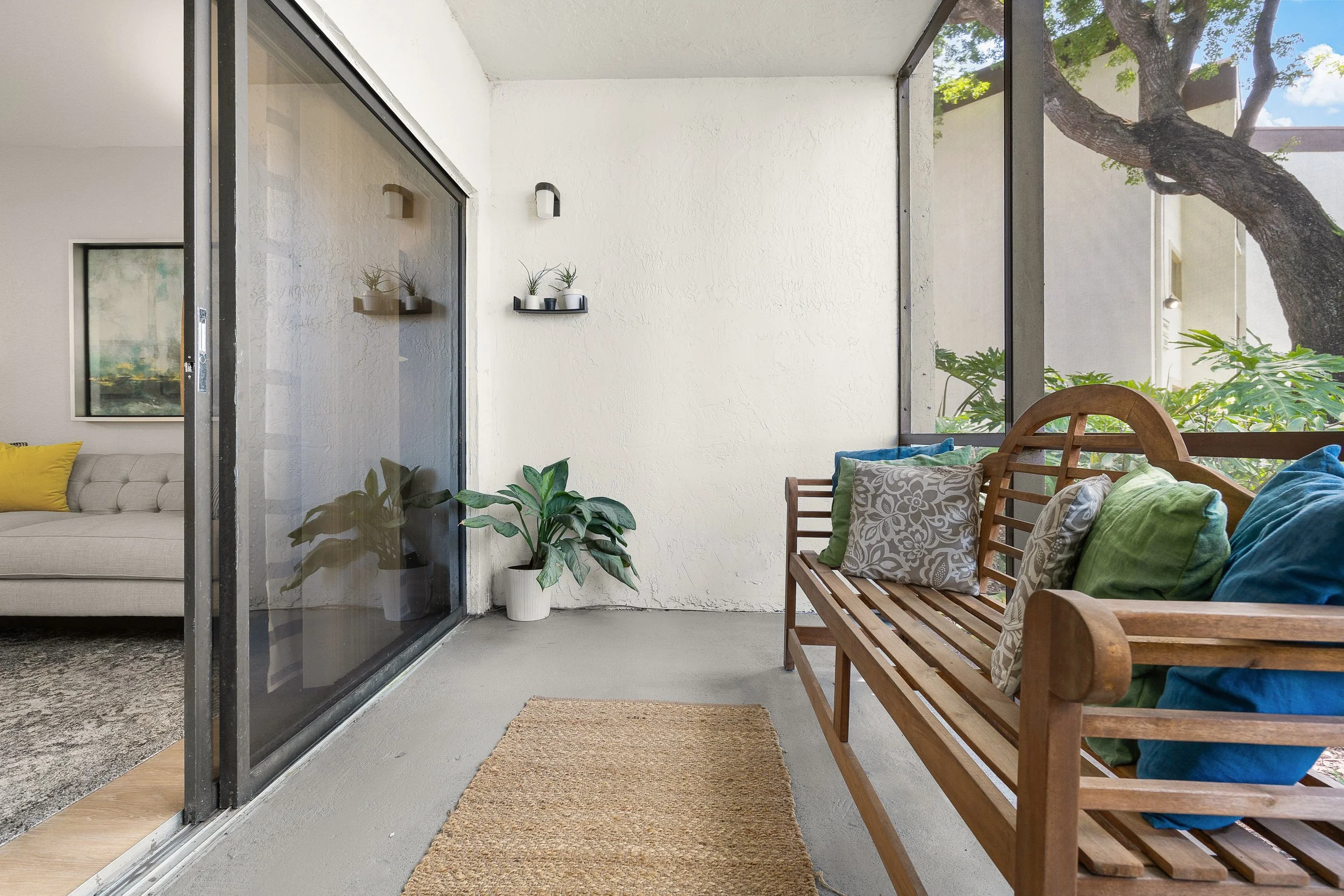 View of a cozy enclosed patio with a wooden bench with colorful pillows, two potted plants, and a sliding glass door leading into a living room with a beige sofa and yellow pillow, with trees visible outside the screened enclosure.