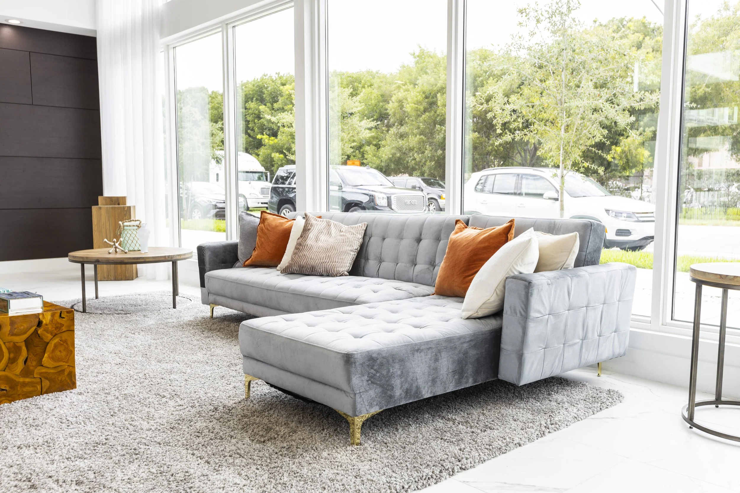Bright living room with a large gray sectional sofa with multicolored pillows, large windows showing parked cars and green trees outside, a round wooden side table, a textured area rug, and light-colored walls and floors.