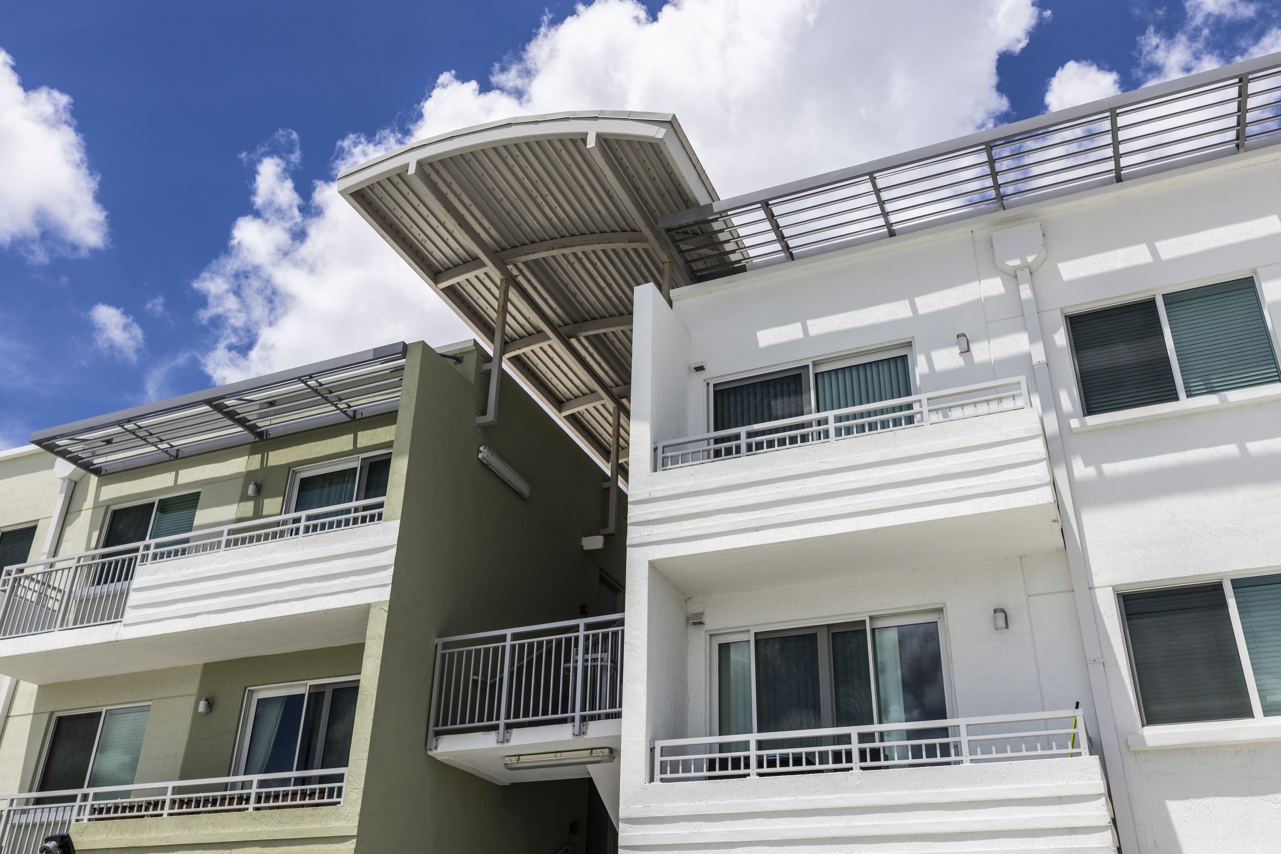 Exterior view of a modern apartment building with balconies, white and green walls, and a blue sky with clouds overhead.
