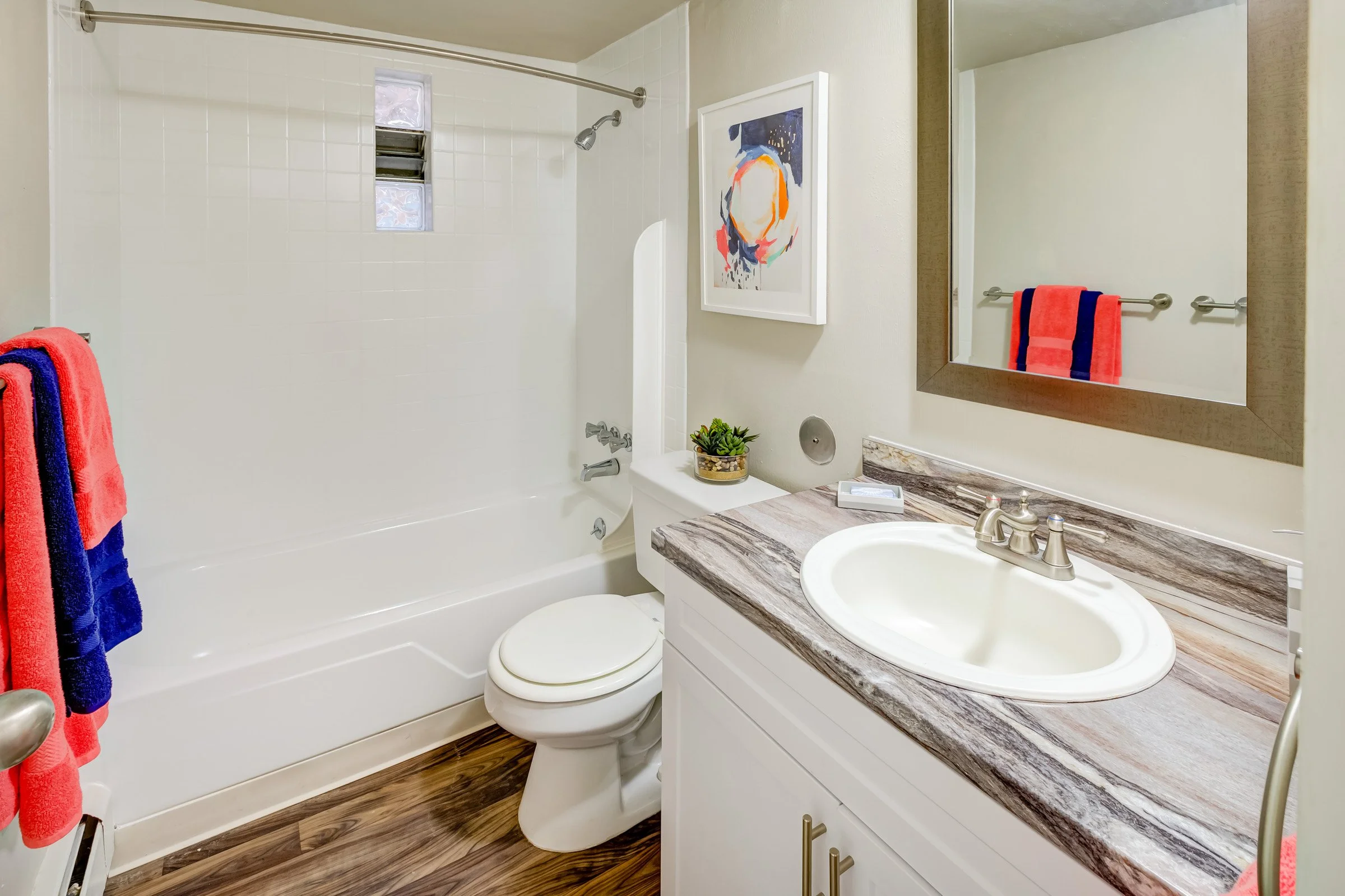 Bathroom with a bathtub/shower, toilet, and a vanity with a sink and mirror. There are colorful towels, a framed artwork, and a small potted plant on the countertop. The floor is wood-style.