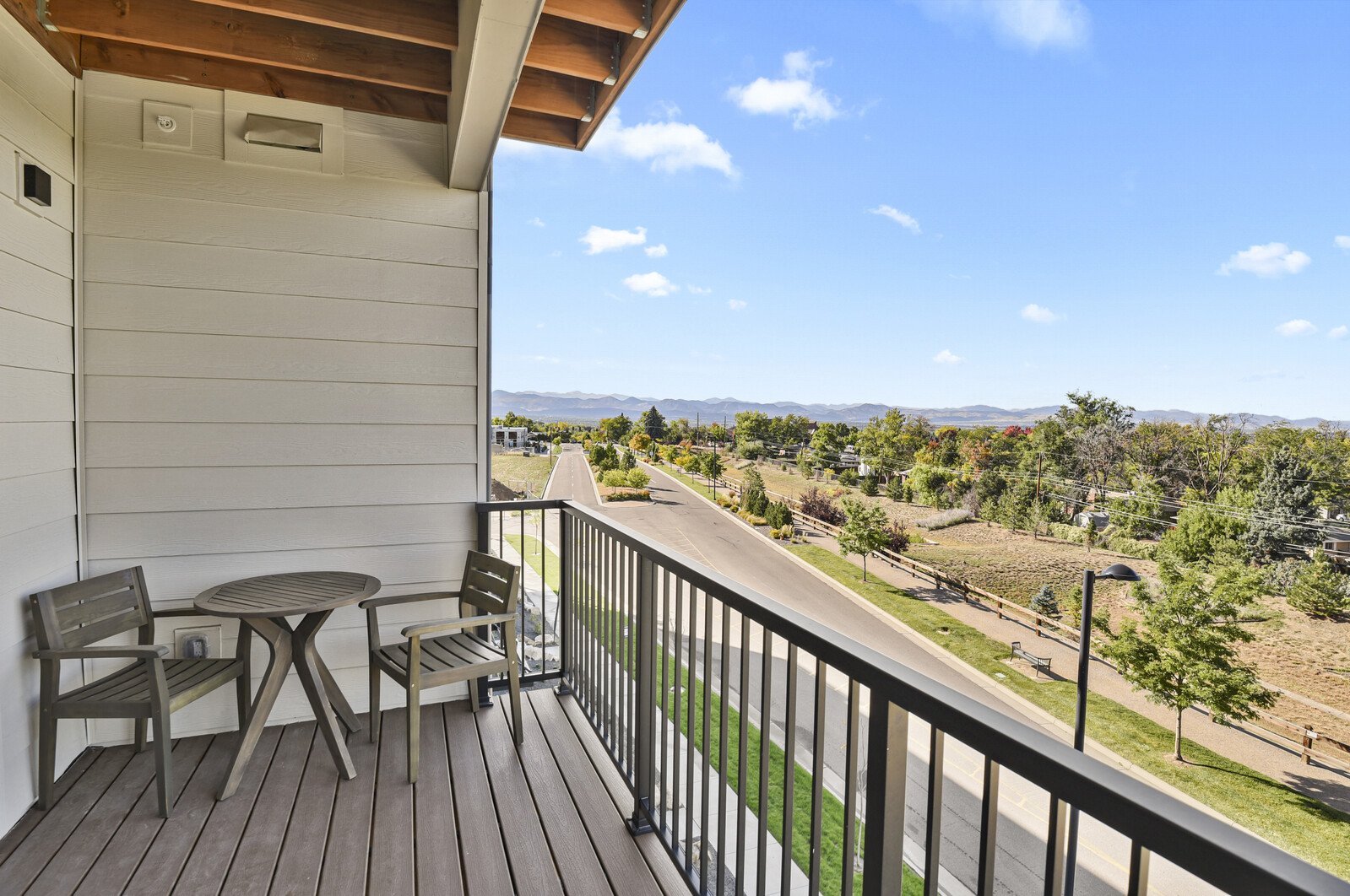 View from a balcony showing a suburban street lined with trees, houses, and distant mountains under a partly cloudy blue sky.
