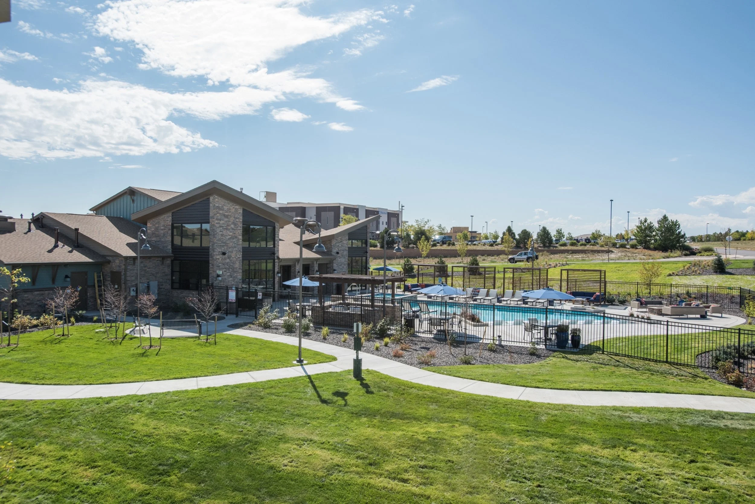 A residential apartment complex with a swimming pool, surrounded by a black metal fence, outdoor seating with umbrellas, a well-maintained lawn, concrete walkways, and a clear blue sky.