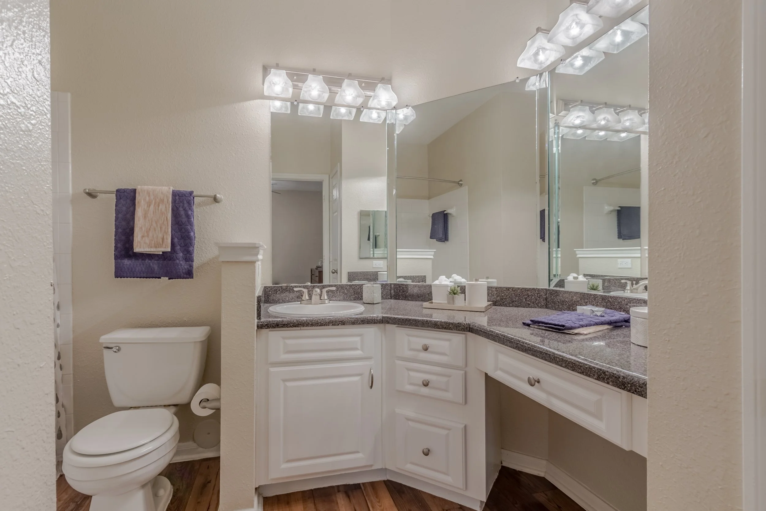 Bathroom with a vanity, large mirror, and white cabinets. Towel rack with purple and beige towels, and toilet. Granite countertop with decorative items and purple towels.